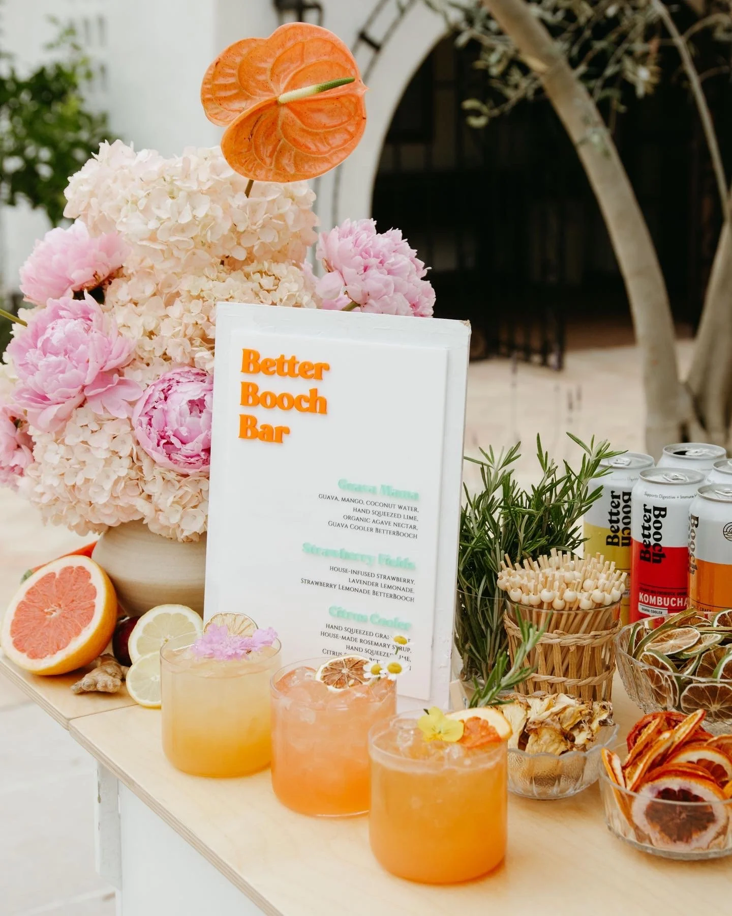 A display of pink and white hydrangeas with an orange anthurium flower, sliced citrus fruits including grapefruit, lemon, and lime, and several colorful drinks with ice. There is a menu sign with the text "Better Booch Bar" and ingredients listed. Th