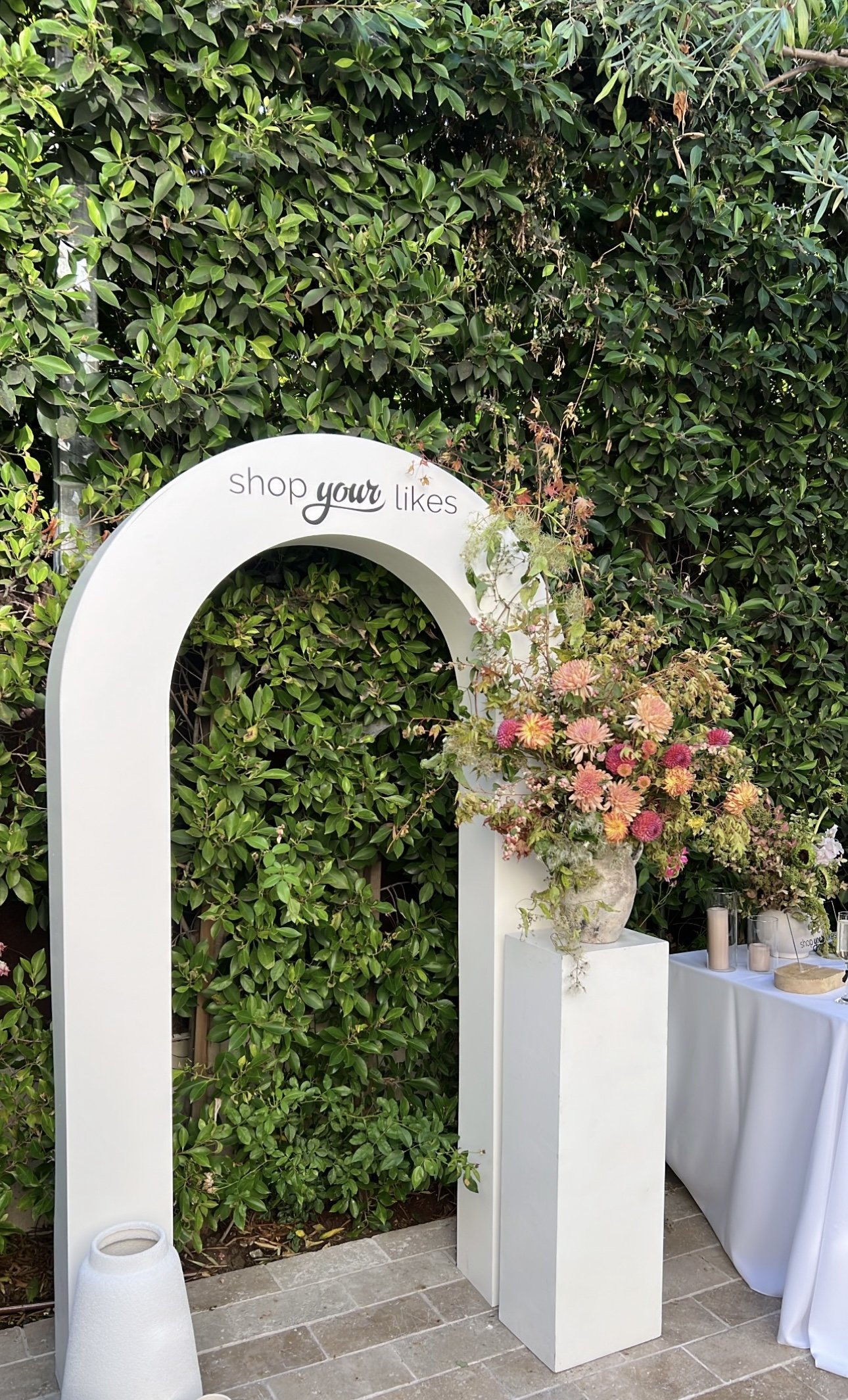 Decorative white arch  backdrop with the vinyl phrase "shop your likes" at the top, next to a large floral arrangement in a white vase, set against a green leafy background and pedastols.