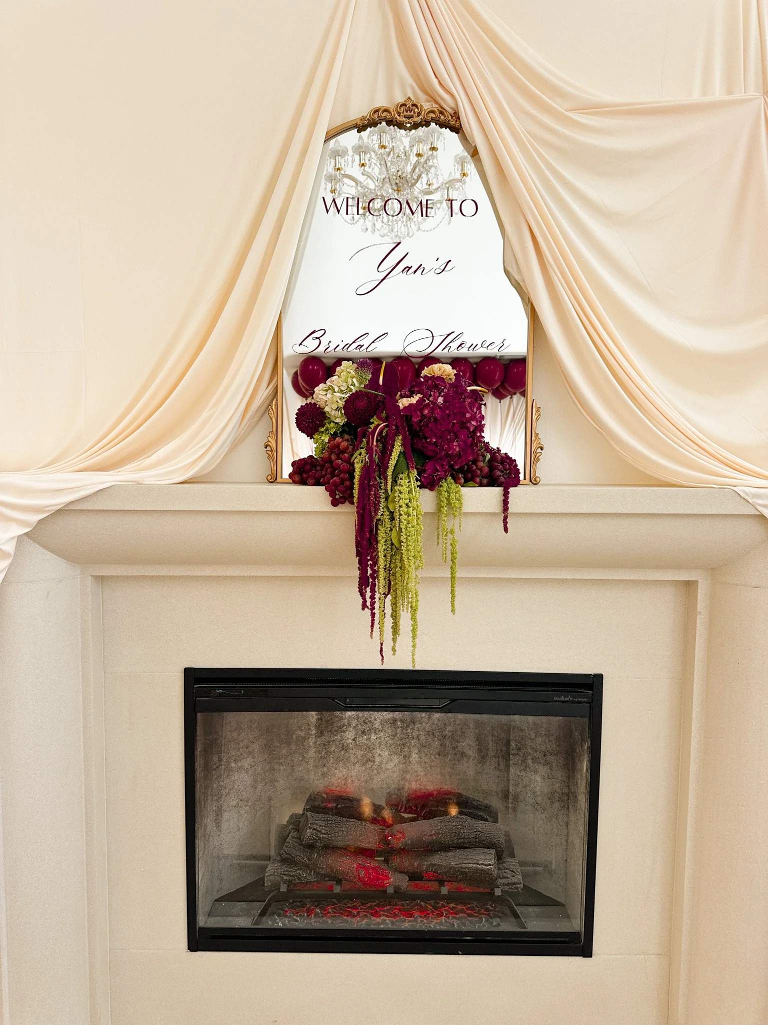 Fireplace with artificial logs and glowing embers beneath a mantel decorated with a floral arrangement of purple and green flowers, with draped cream-colored curtains and a mirror with a chandelier reflected in it.