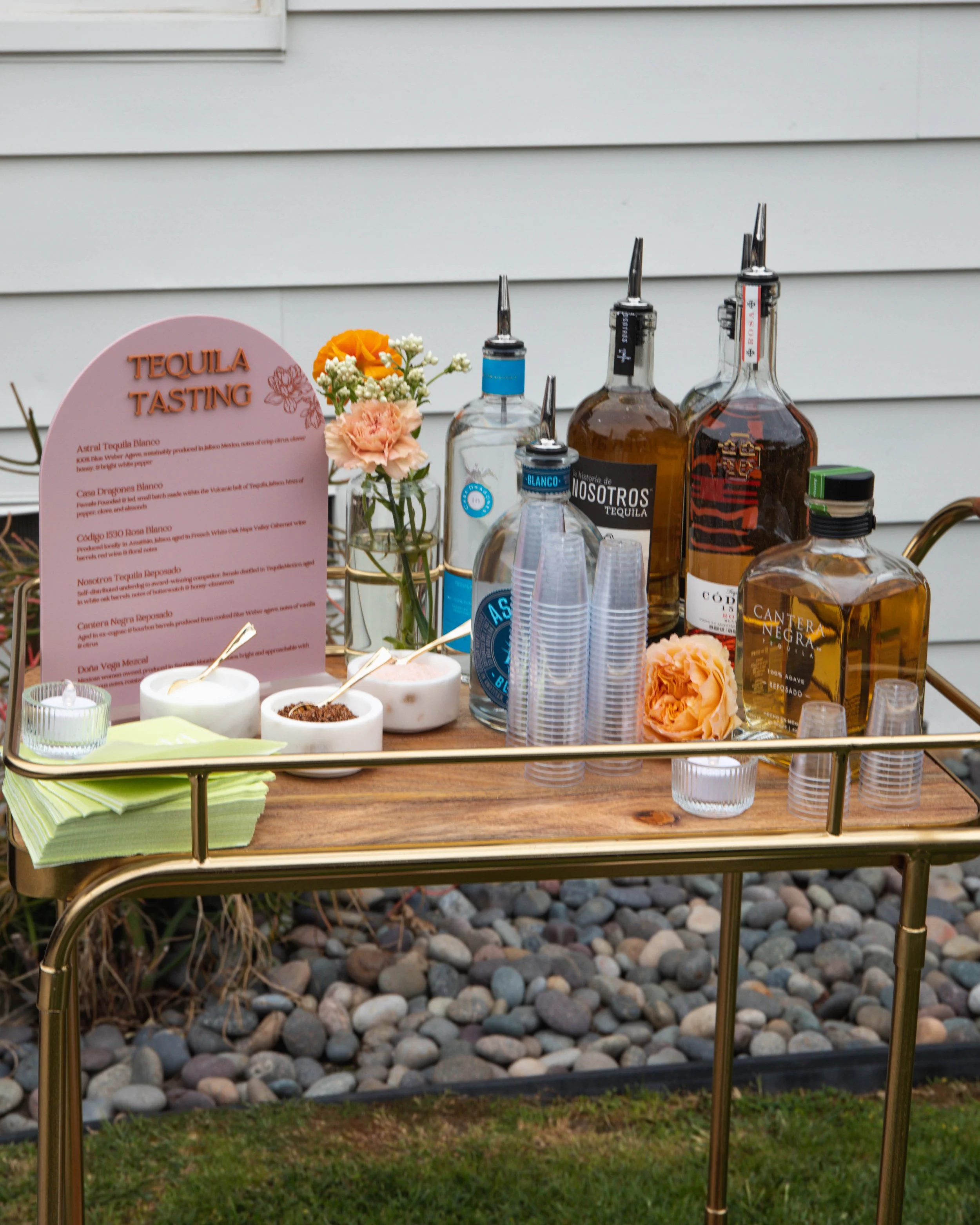 A table set up outdoors with bottles of alcohol, a small bouquet of flowers, and a menu for a tequila tasting event, with a backdrop of q-siding and a rocky ground.