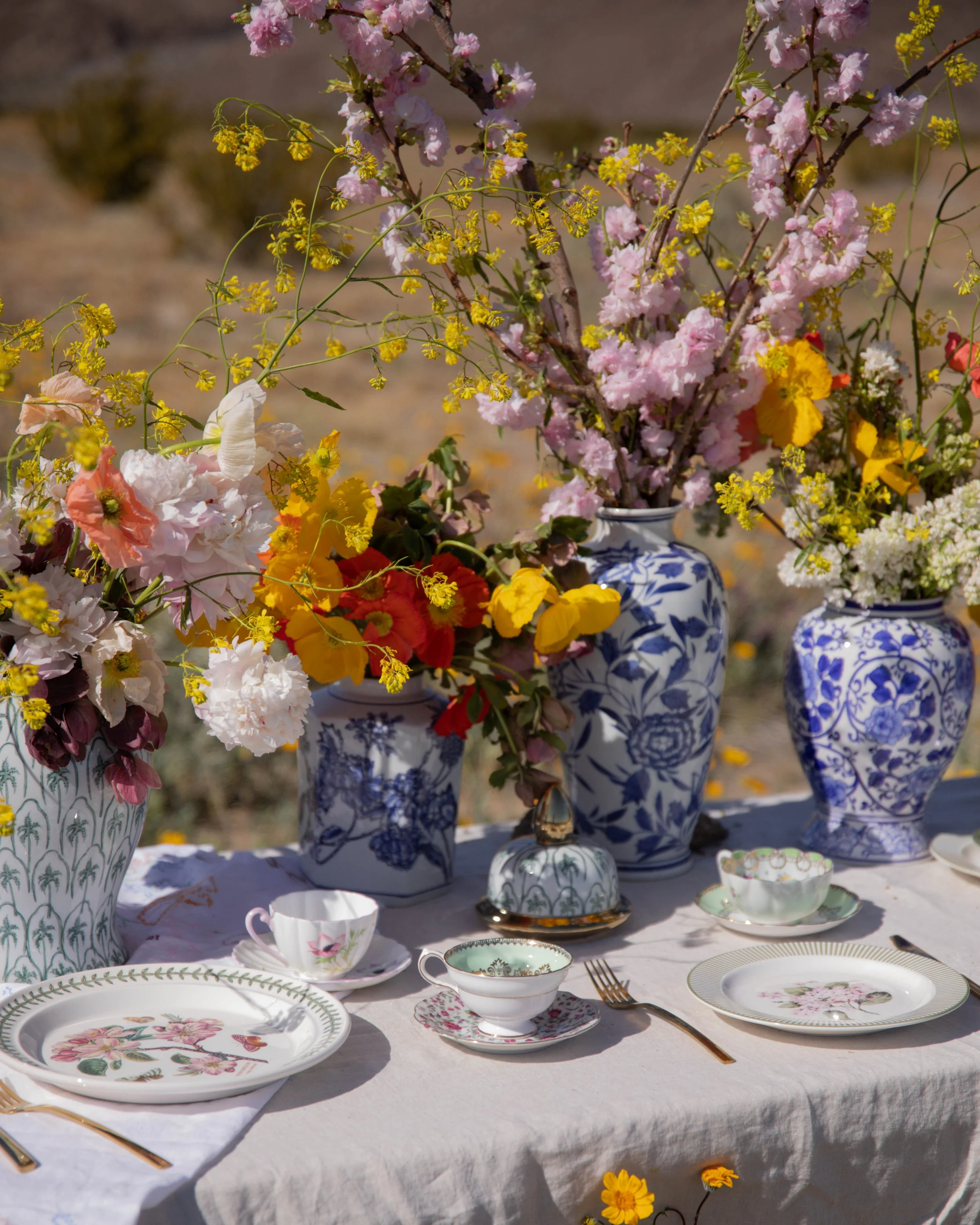 Vase of pink and white flowers, flowers in bowls, and elegant porcelain plates on a table with a white tablecloth, set outdoors during daytime.