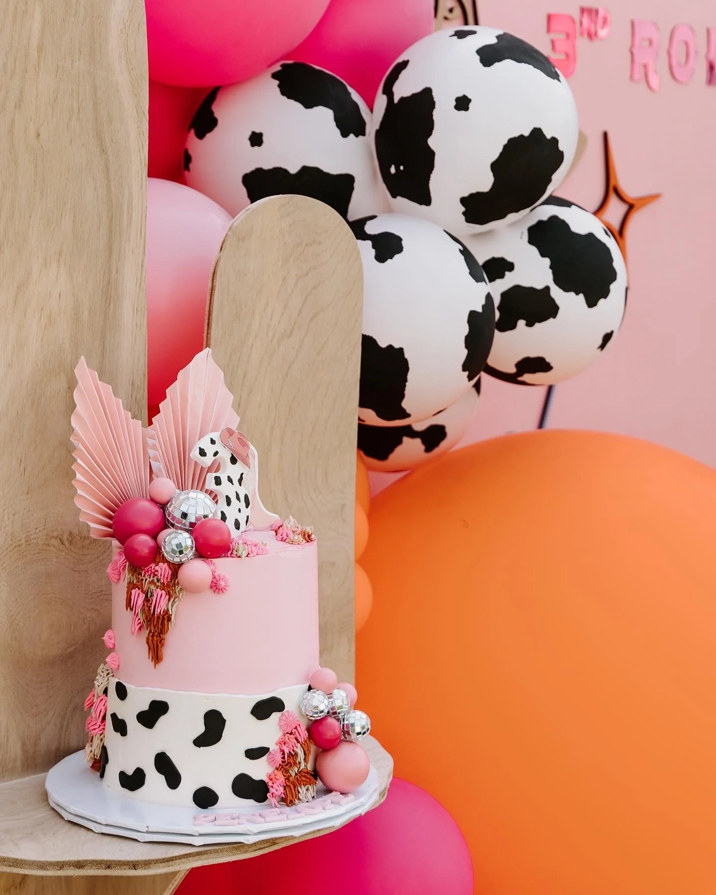 Pink and black spotted cow-themed cake decorated with pink, silver, and black balls, pink paper fans, and cow print accents, with a background featuring pink and orange balloons.