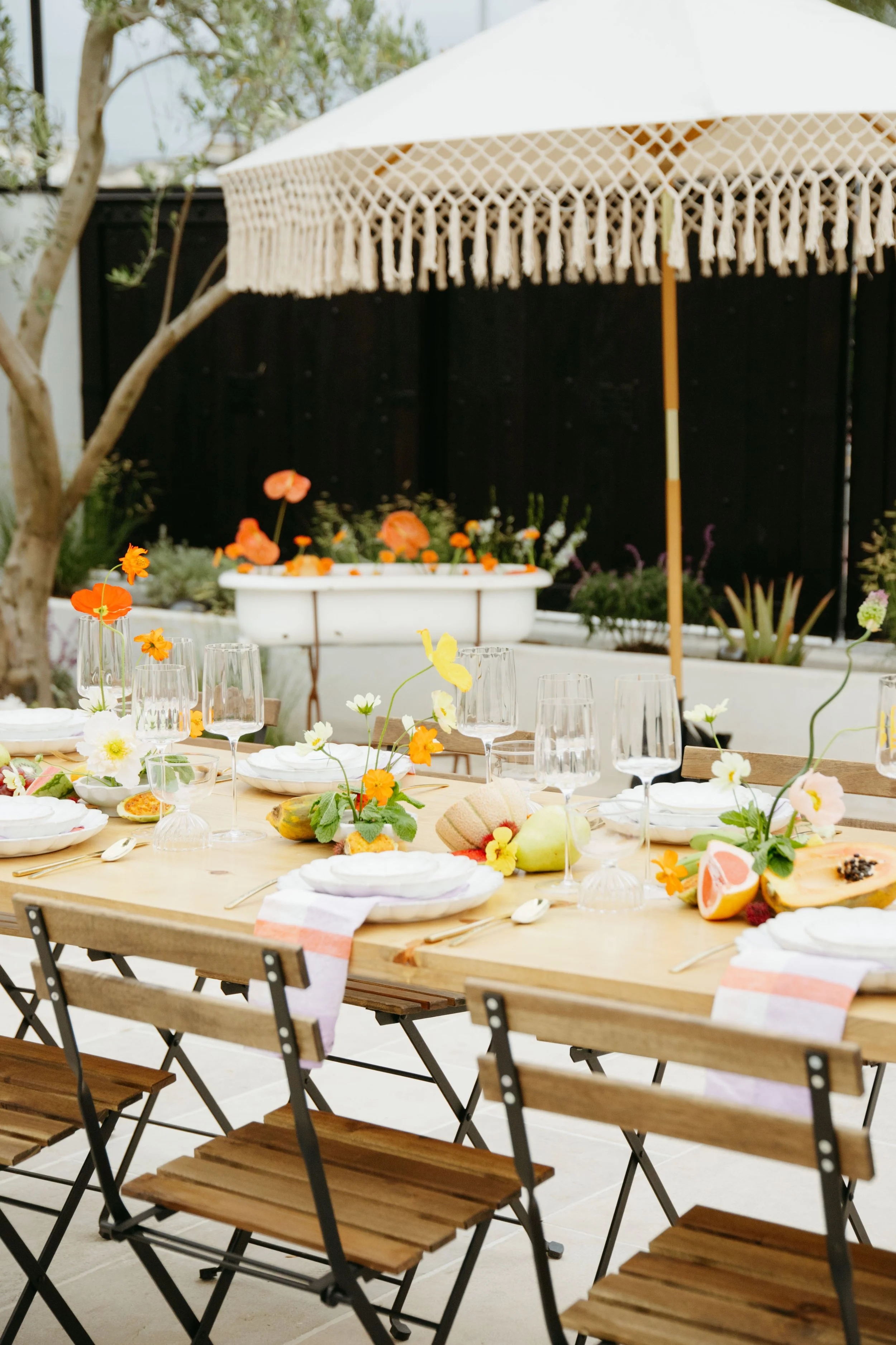 Outdoor dining table decorated with flowers and food. In the background, there is a large white umbrella and a black fence, with trees visible.