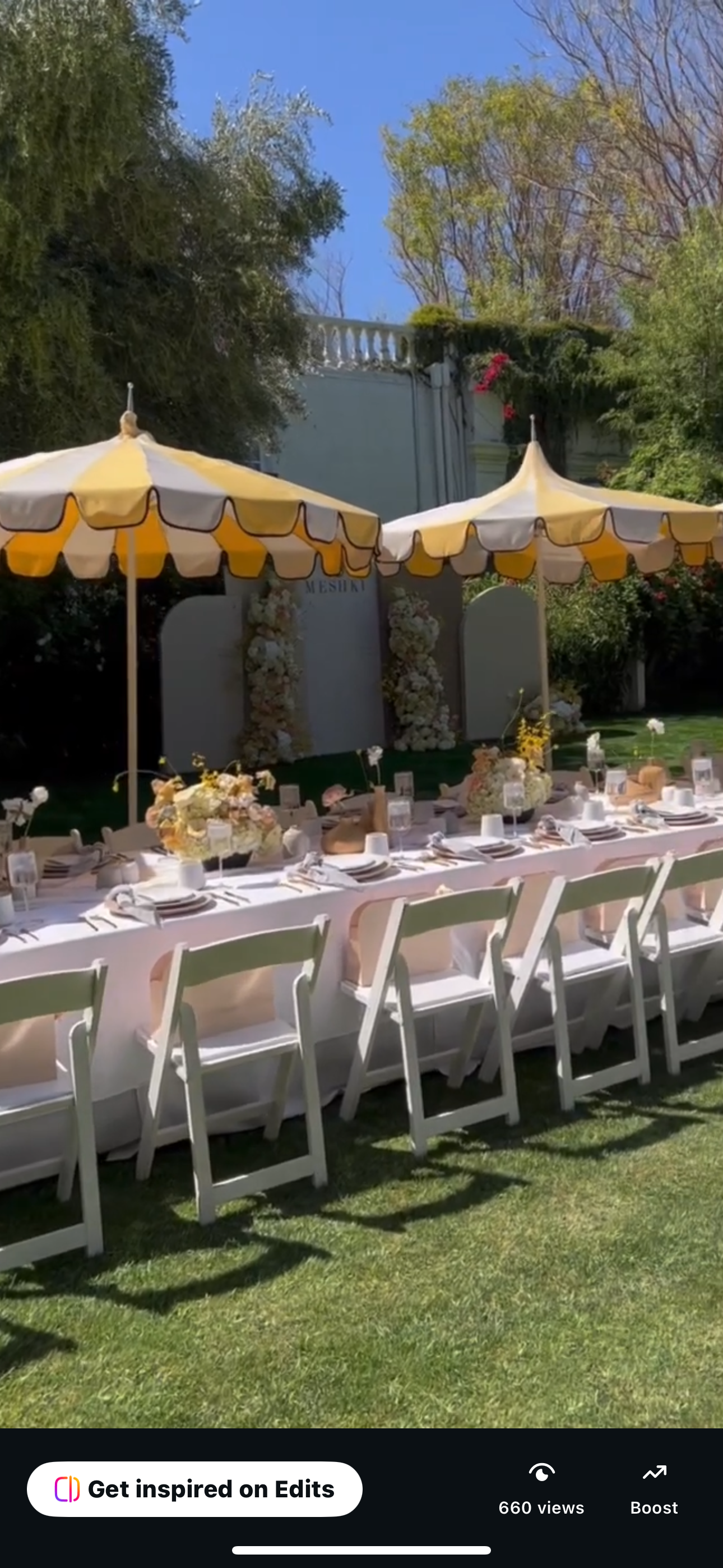 Outdoor wedding reception setup with a long table covered with a white tablecloth, decorated with floral centerpieces, surrounded by white chairs, under yellow and white umbrellas on a grassy area, with trees and a building in the background.