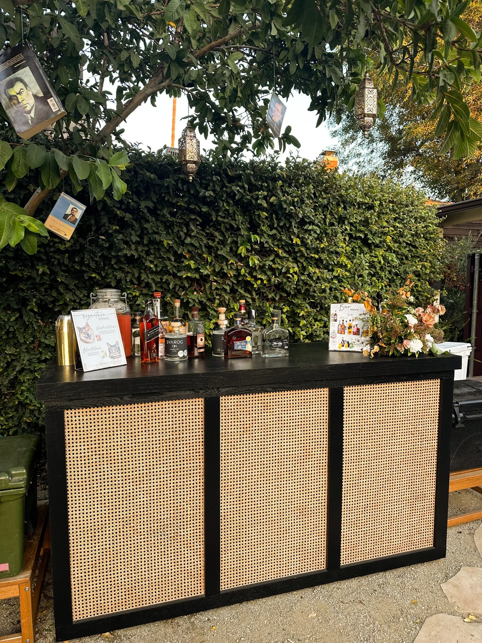 An outdoor bar with a black frame and rattan panels, situated in front of a lush green hedge and wooded area, displaying bottles of alcohol, cocktail ingredients, and decorative signs.