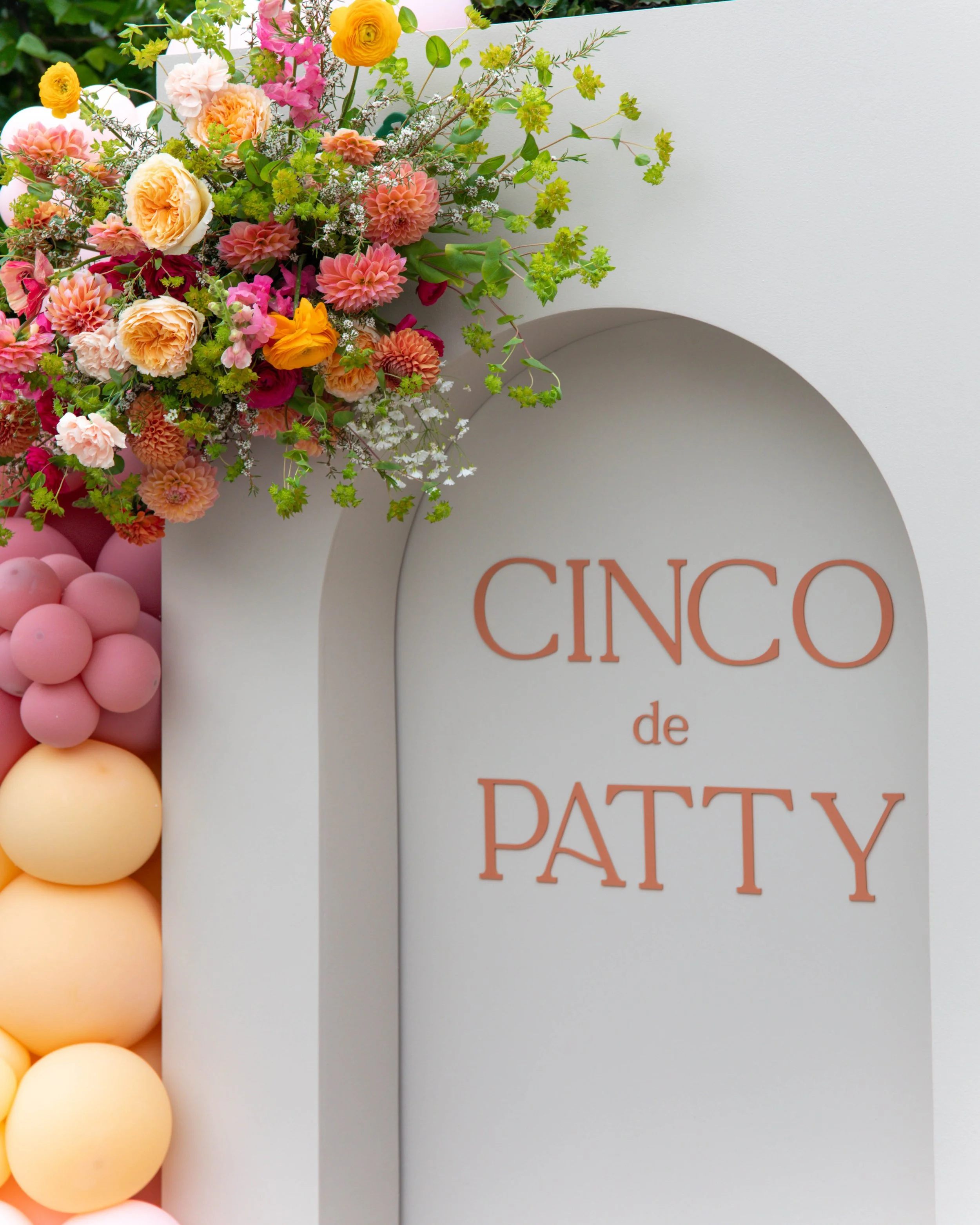 Colorful flower arrangement with pink, peach, yellow, and white flowers, along with greenery, displayed next to a white circular sign with orange text that partially reads 'Cinco de...' and a balloon decoration with shades of pink and peach balls.