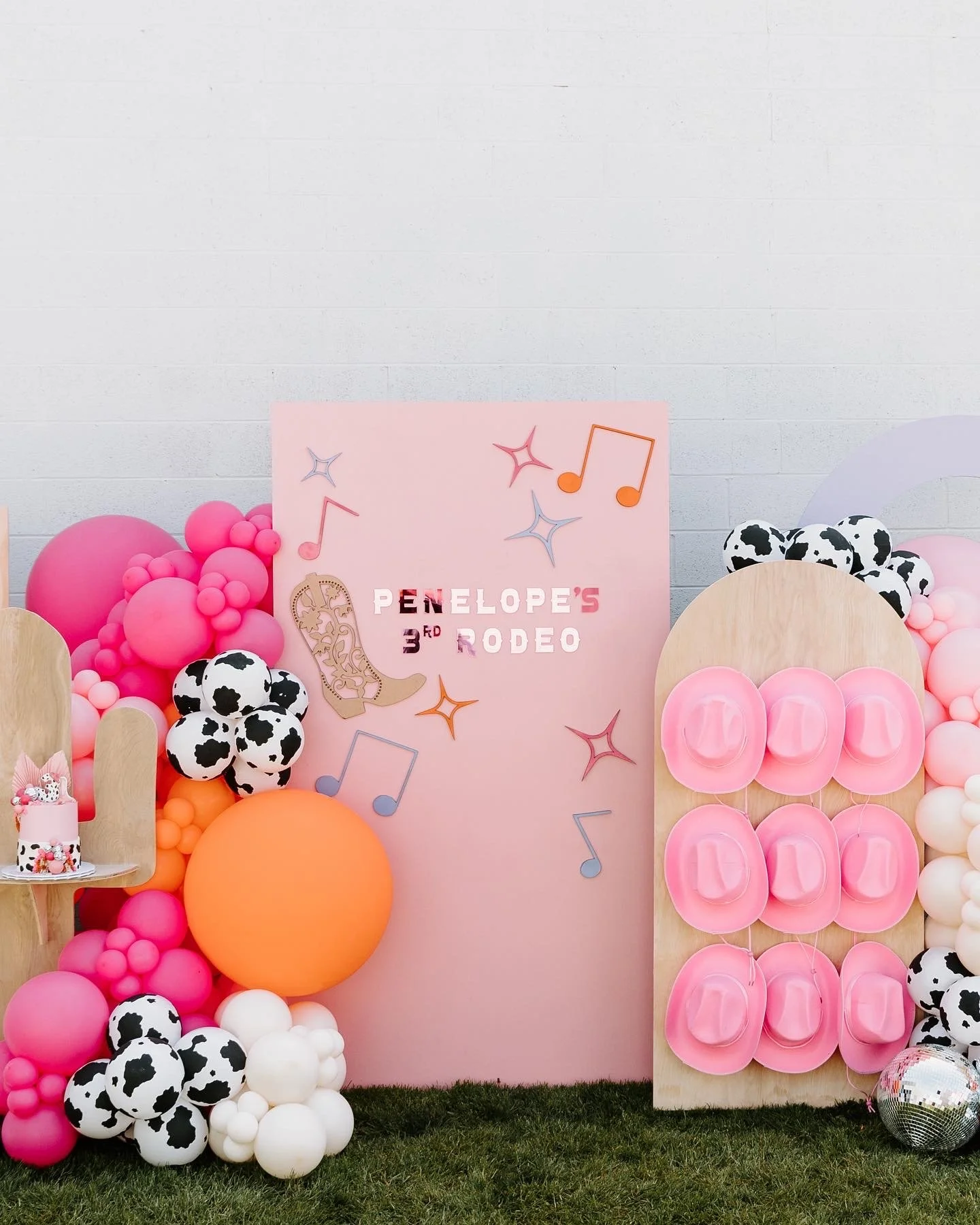 Party backdrop with pink, white, orange, and black-and-white balloons, a pink sign with music notes and the text "Penelope's 3rd Rodeo," and a small birthday cake on a wooden chair.