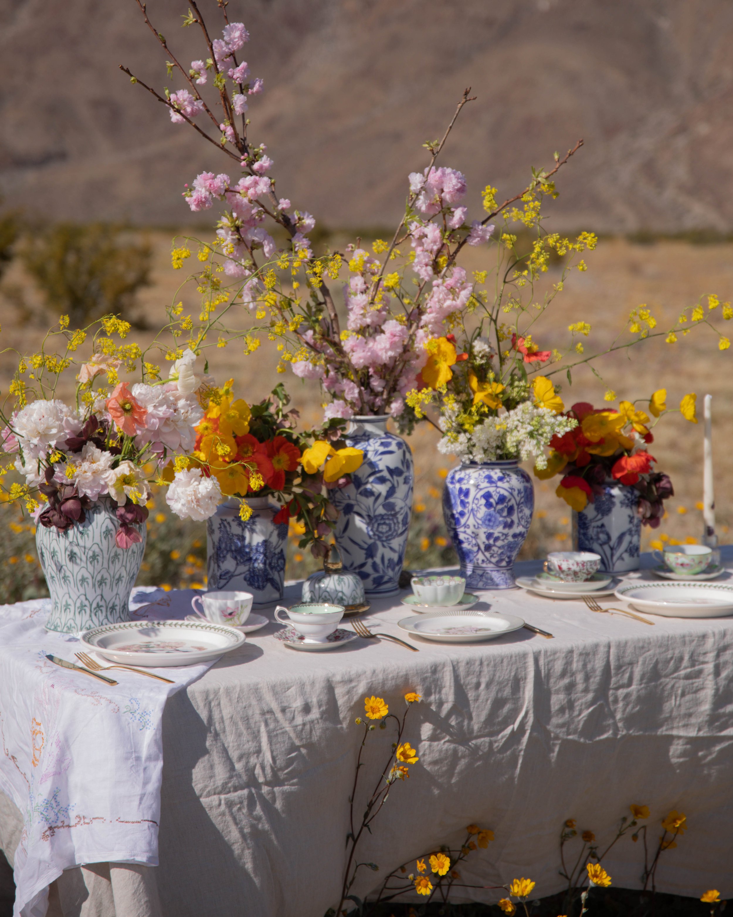A table set with floral china vases filled with pink, yellow, white, and red flowers, and set with plates, cups, and gold utensils, outdoors with desert background and yellow flowers at the base of the table.