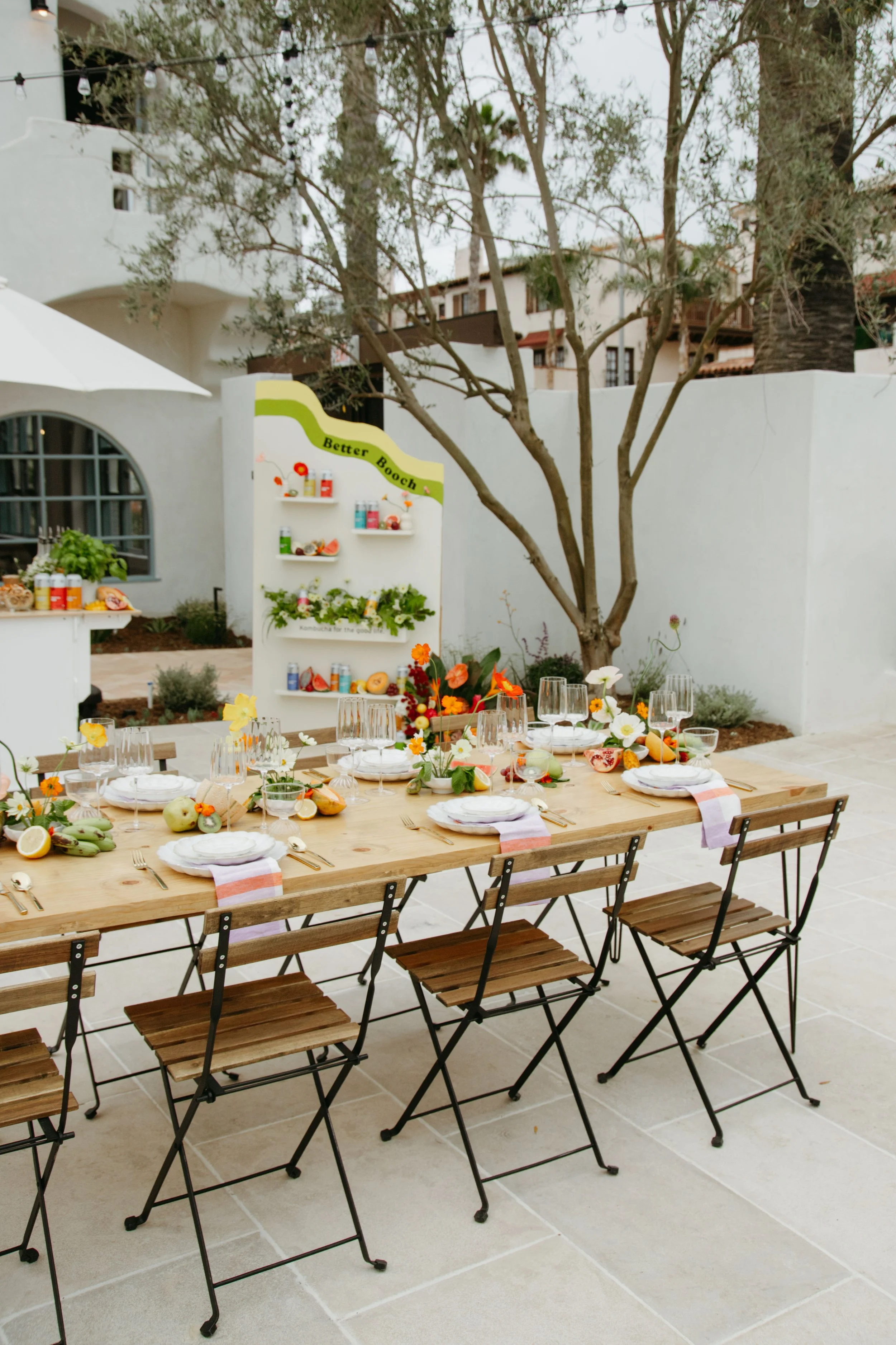 Outdoor dining setup with a long wooden table, chairs, place settings, and a floral centerpiece, with a mural and trees in the background.