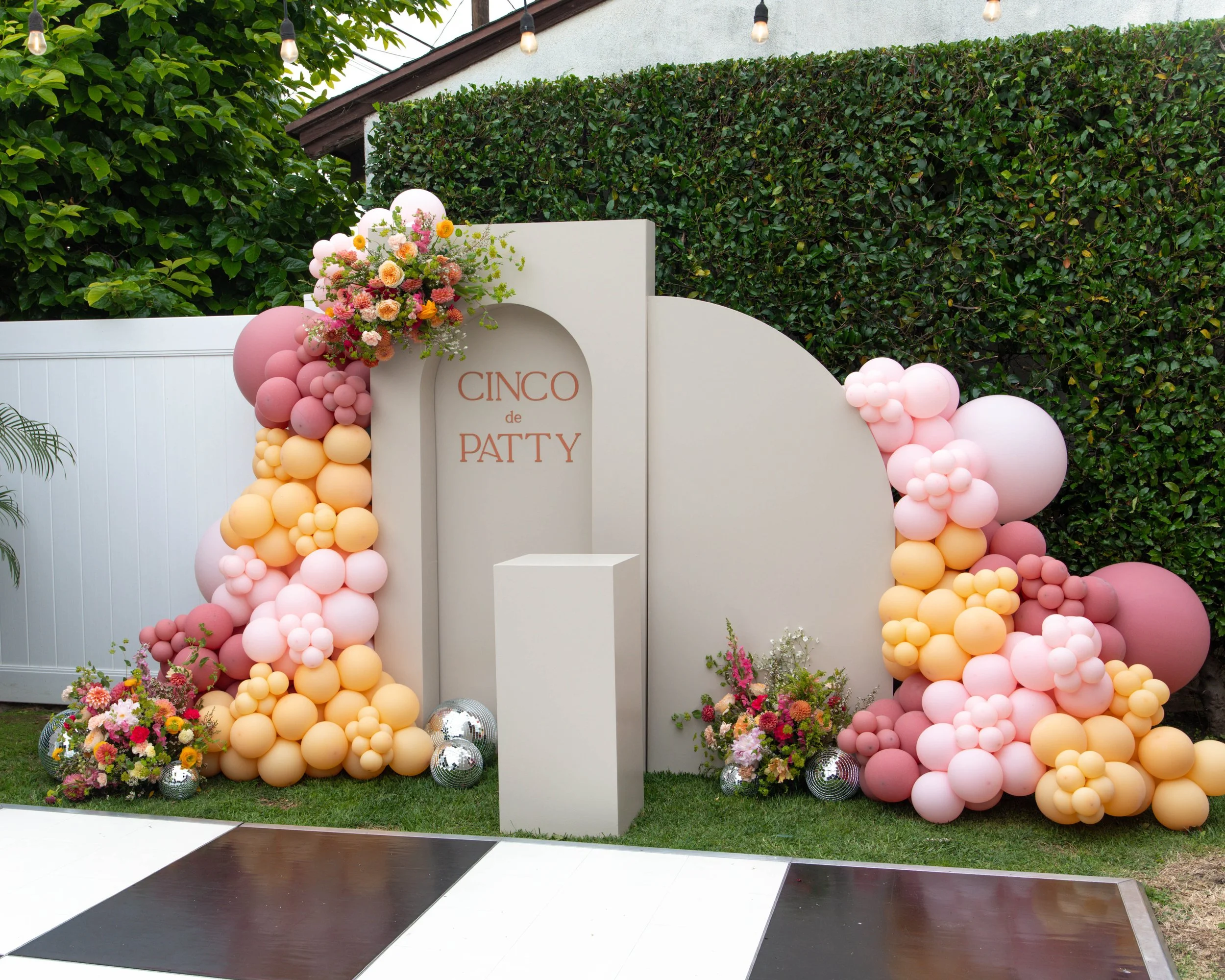 Event backdrop with pink, peach, and white balloons, floral arrangements, and a sign that says "Cinco de Patty" amid greenery and string lights.