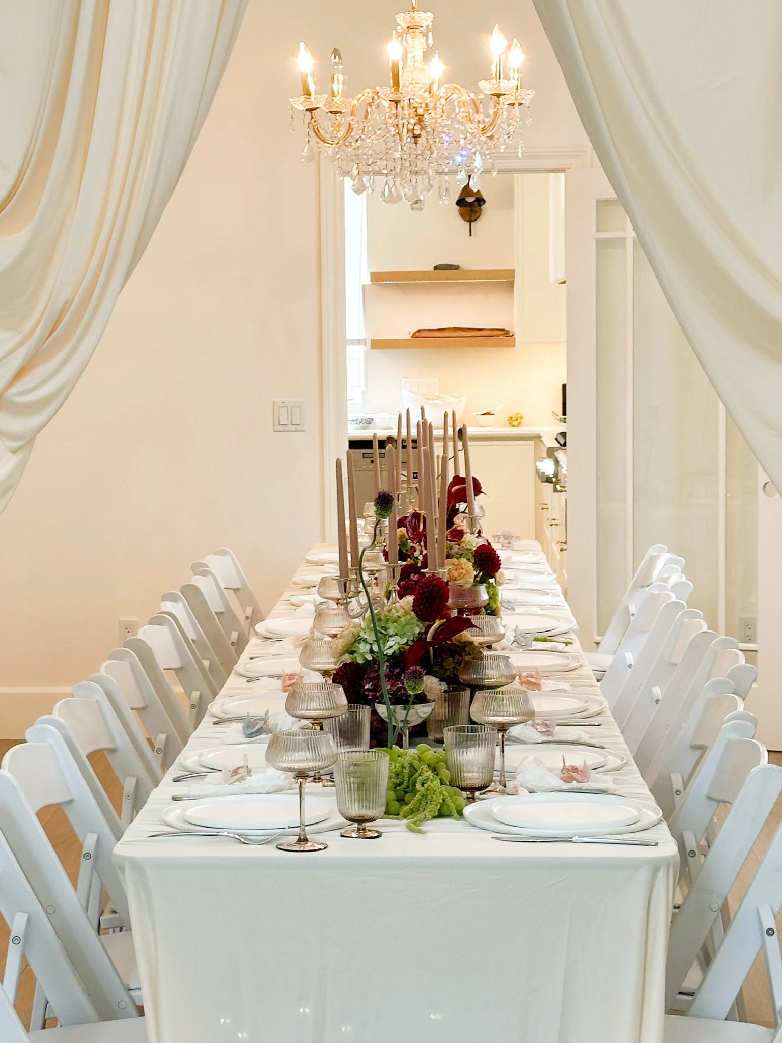 A long dining table decorated with a white tablecloth, floral centerpieces, candles, and individual place settings, set up for a formal event in a bright room with a chandelier hanging from the ceiling.