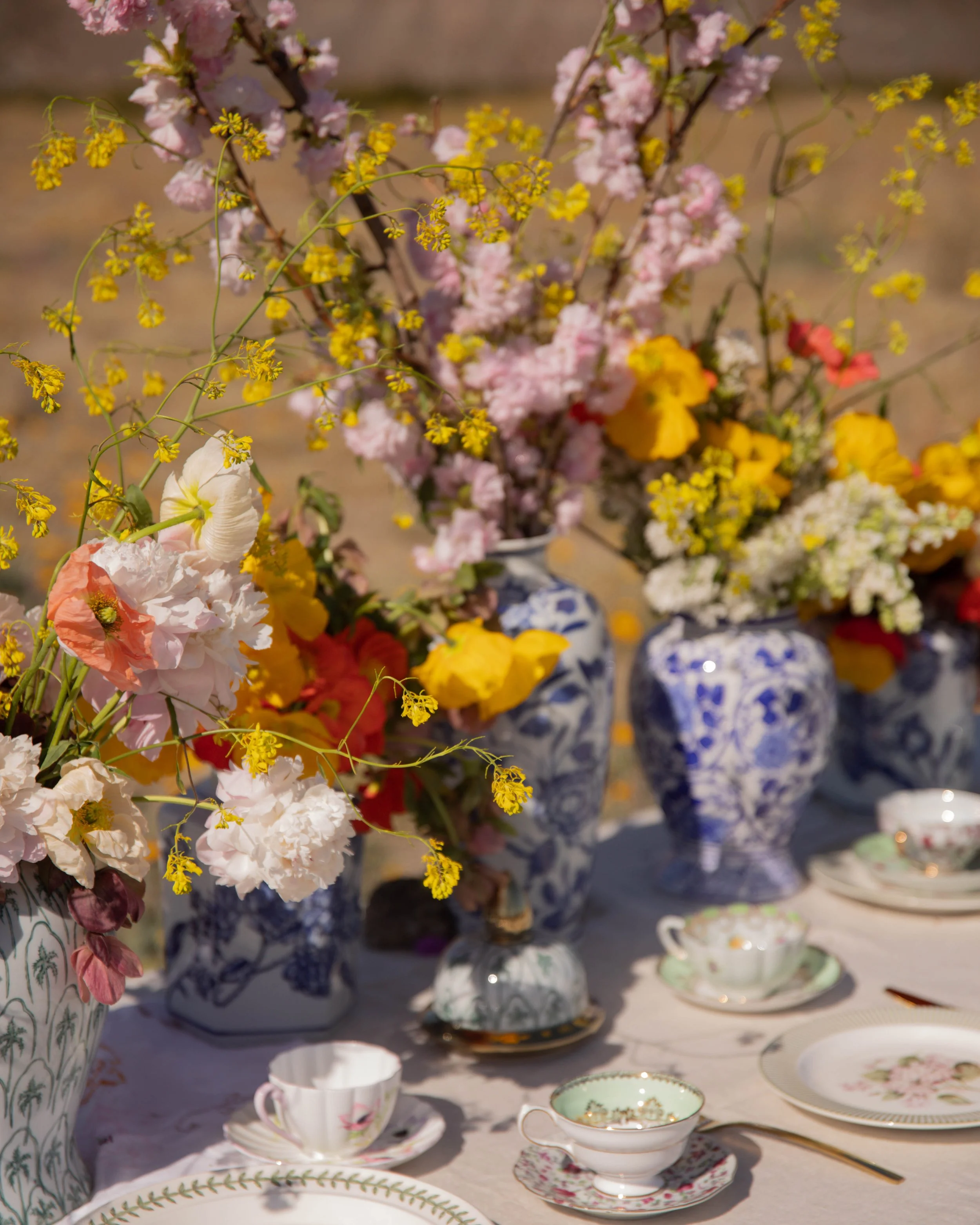 A table filled with vintage teacups, saucers, and plates with floral patterns, set with a large floral arrangement of pink, yellow, and white flowers in decorative vases.