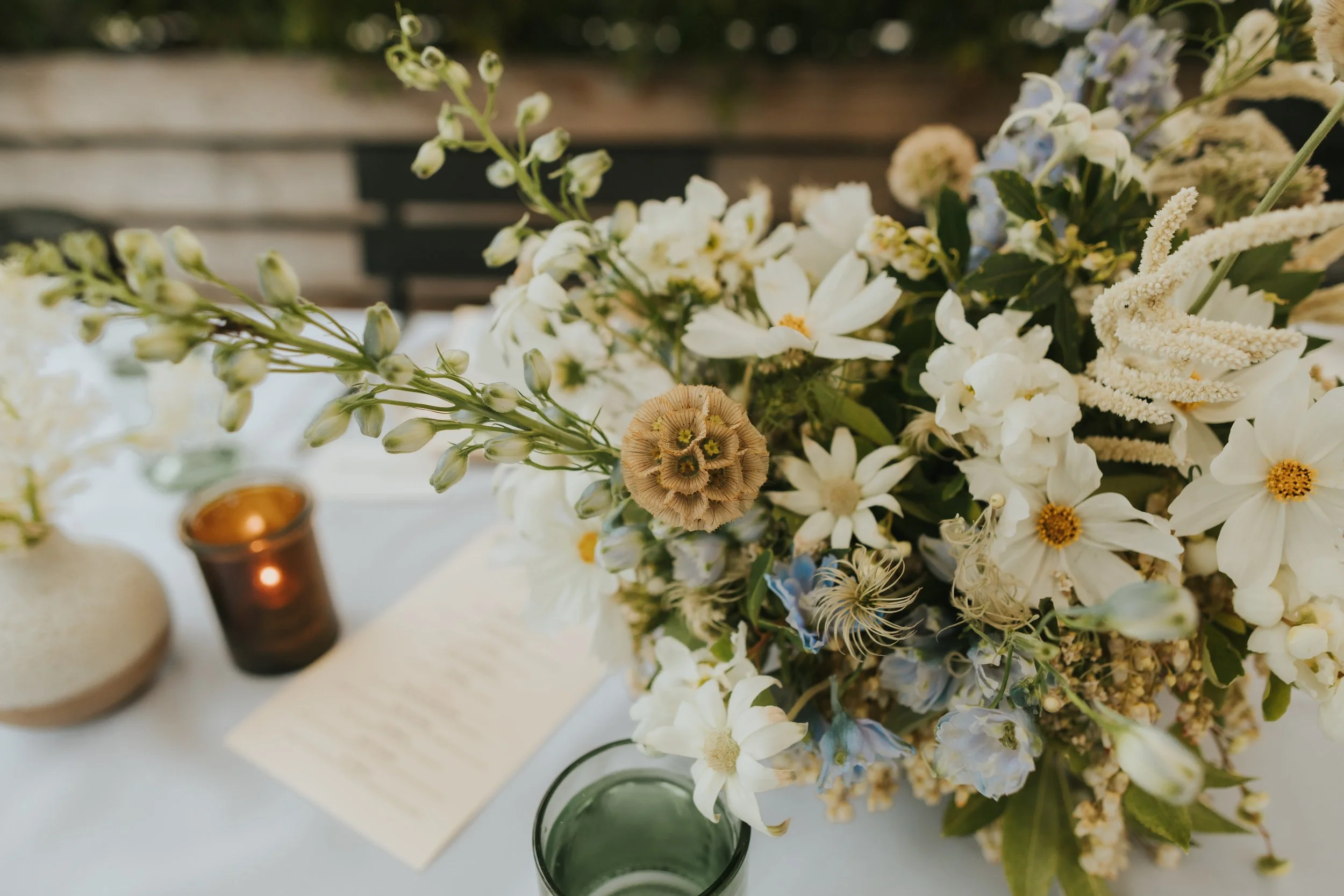 A large bouquet of white and light blue flowers on a table, with candles in small glass holders and a blurred background.