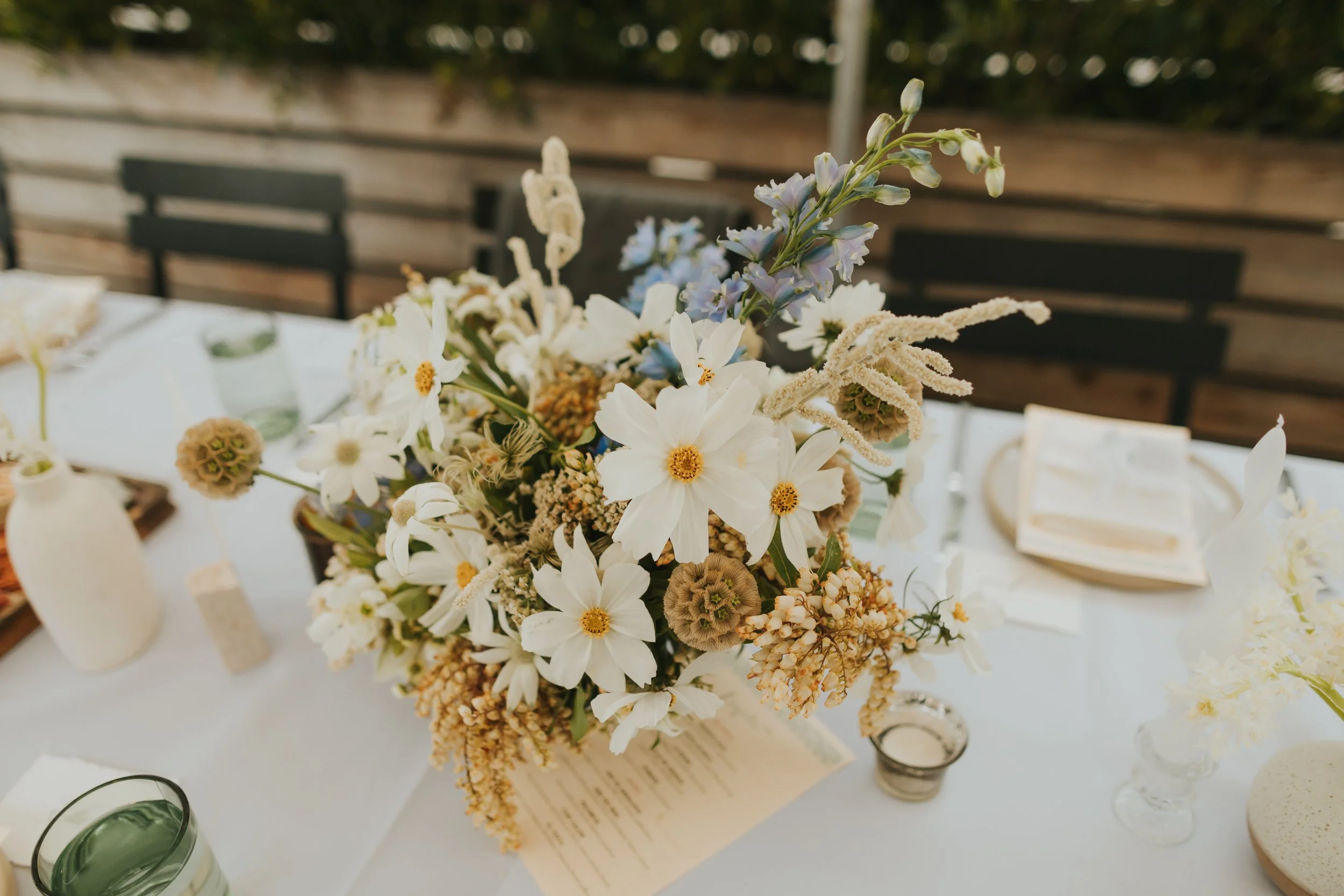 A floral centerpiece on a white tablecloth with votive candles and utensils at an outdoor event.