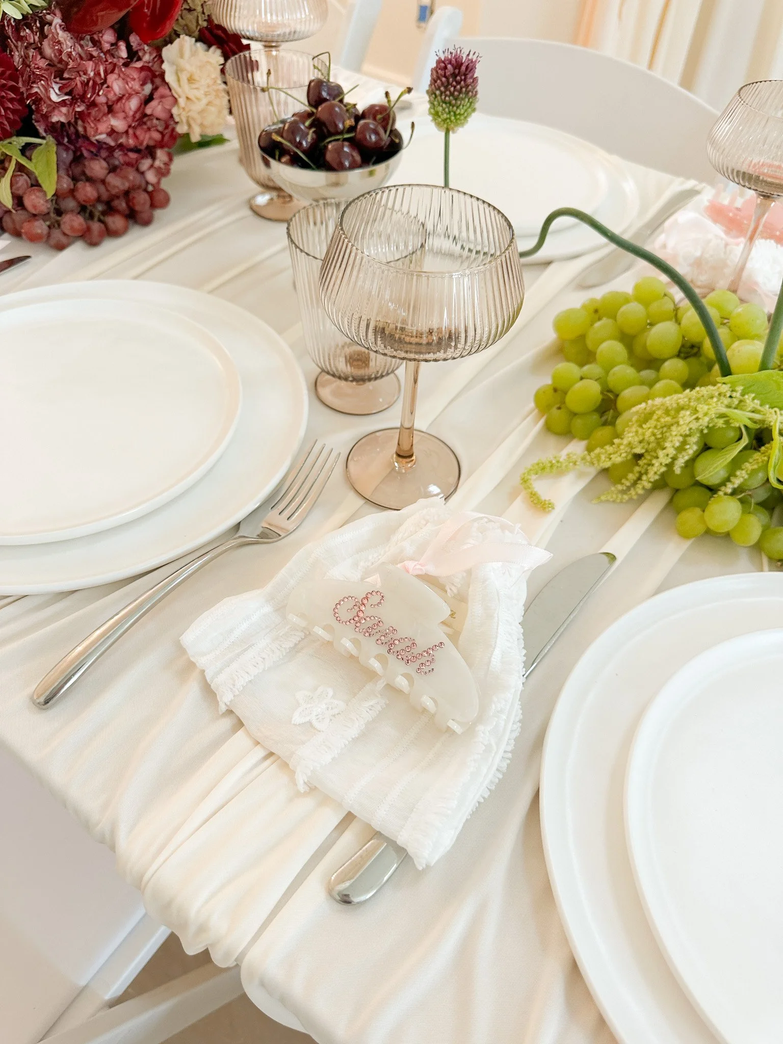 A table decorated for a celebration with a white tablecloth, empty white plates, silverware, wine glasses, and a centerpiece featuring clusters of green and purple grapes, along with a pink flower and a pink decorative item.