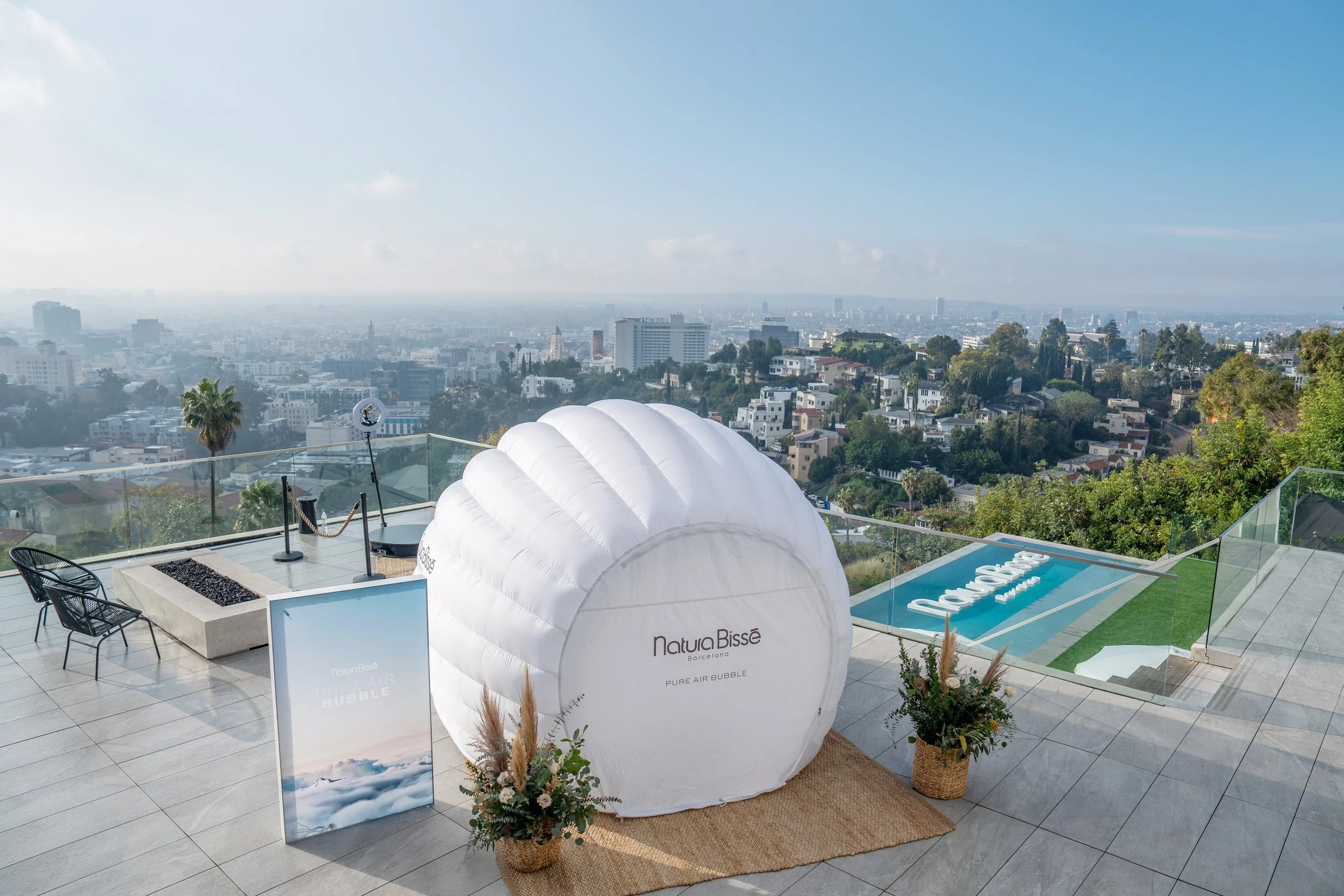A rooftop terrace with a city view, featuring a large white inflatable bubble with 'Natura Bissé' written on it, surrounded by potted plants, chairs, and a glass barrier, under a clear sky.