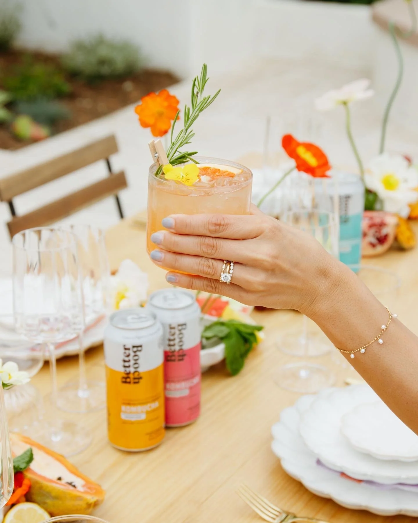 A hand holding a glass of cocktail with orange and yellow flowers garnishing it, surrounded by cans of bubbly water, empty glasses, flowers, and decorative plates on a table at a celebration or gathering.