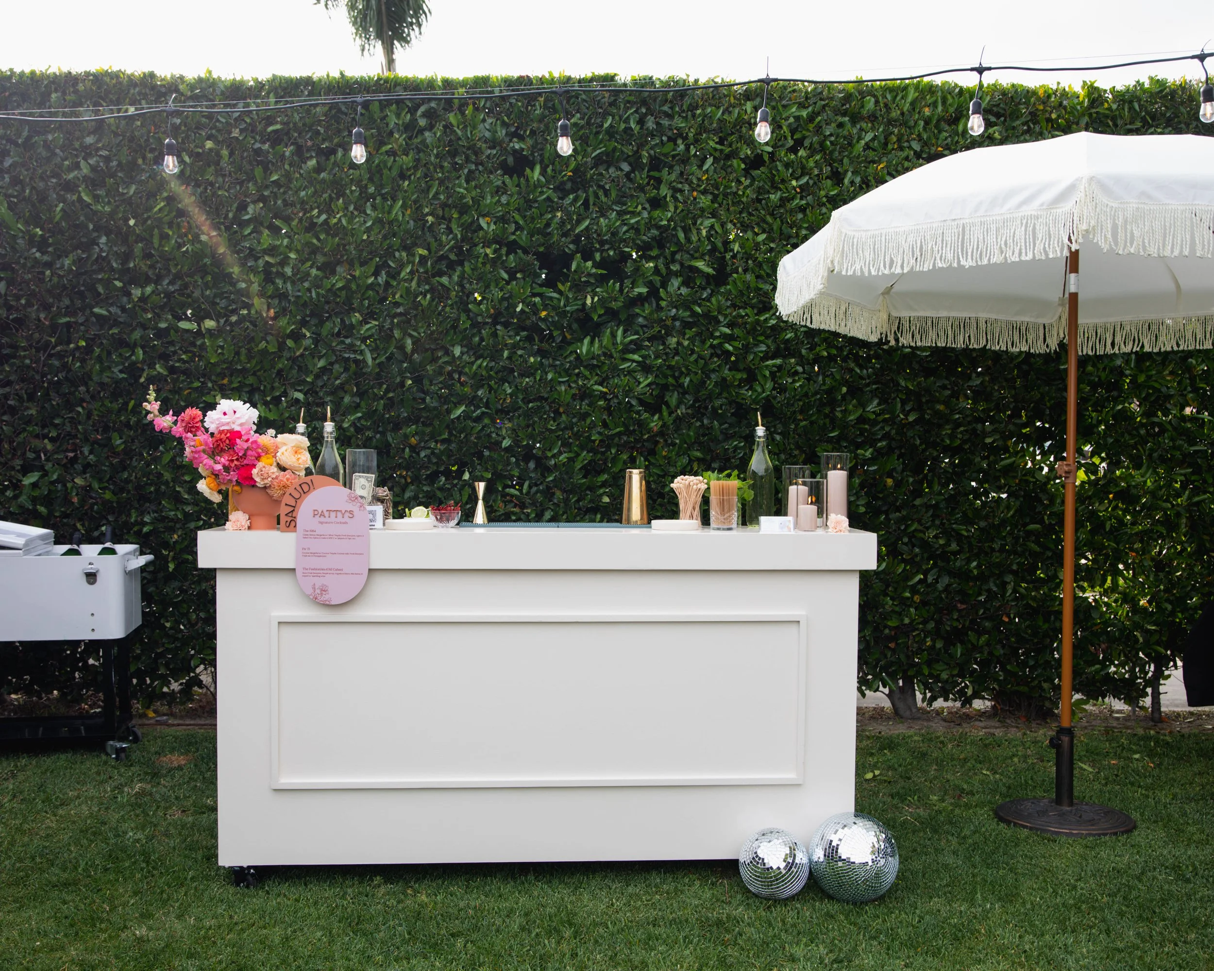 Outdoor event setup with a white bar or table, decorated with flowers, candles, and drink bottles, under a large white canopy umbrella, with string lights hanging above and a grassy ground with disco balls at the base, against a backdrop of tall gree