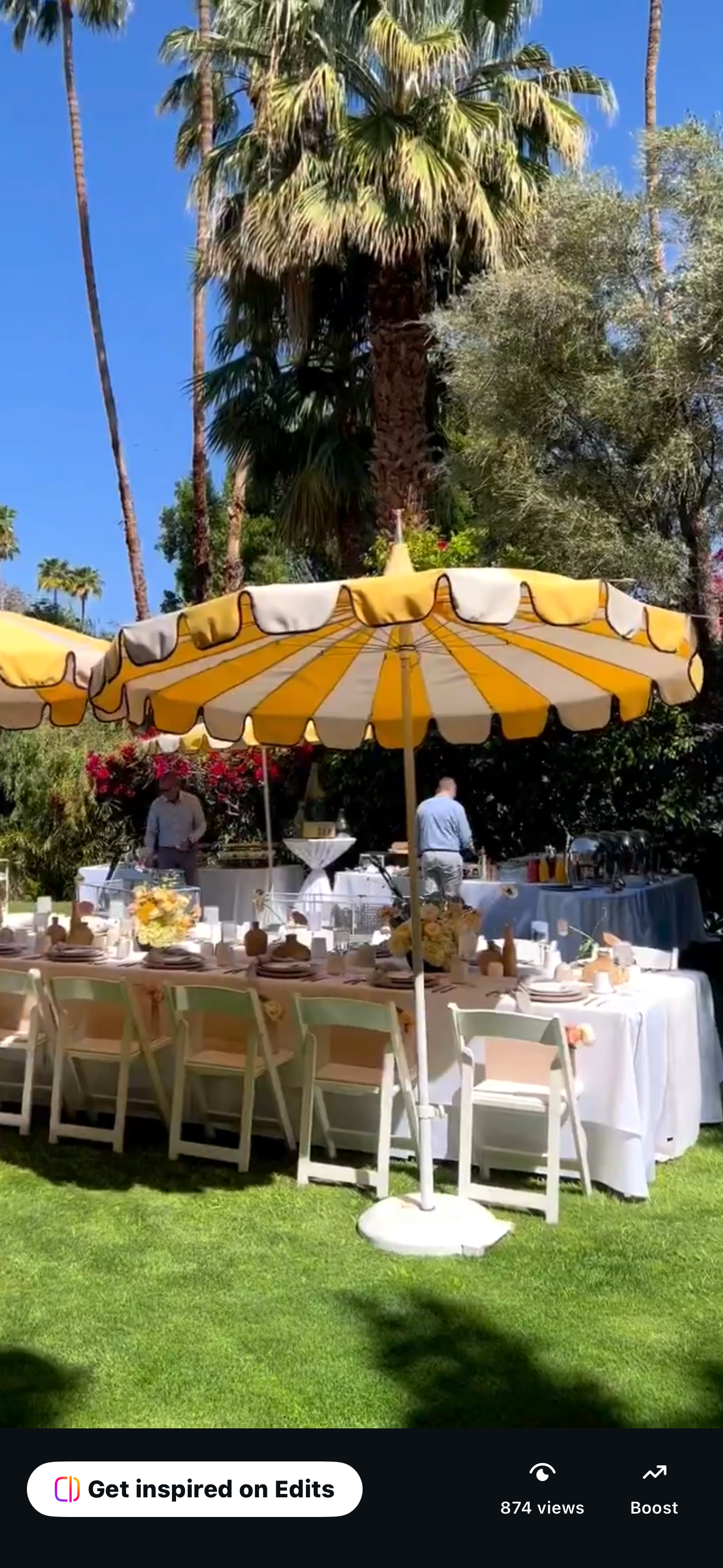 Outdoor garden party setup with a long table, decorated with flowers and tableware, under a yellow and white striped canopy. Palm trees and lush greenery are in the background, with a clear blue sky overhead.