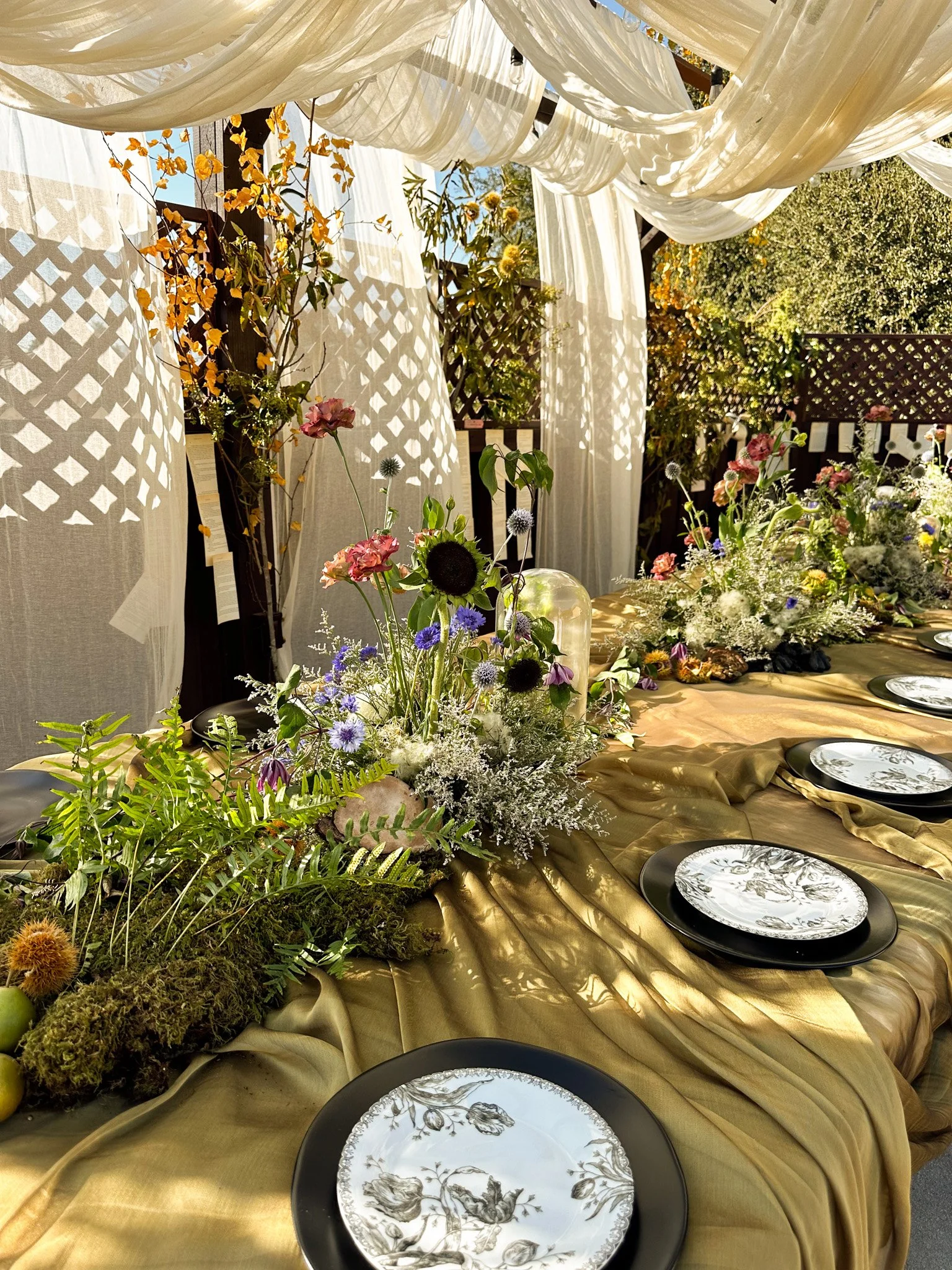 Decorative outdoor table setup with floral arrangements, draped white fabric overhead, and black and white plates on a gold-colored tablecloth.