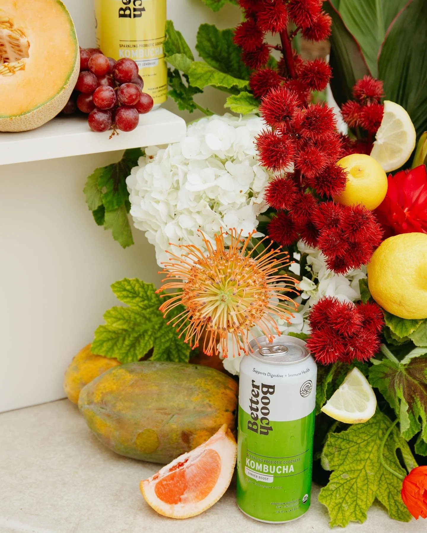 Arranged fruits and flowers including a halved cantaloupe, red grapes, grapefruit slices, red and white flowers, greenery, a soda can, a red flower, and some tropical leaves.
