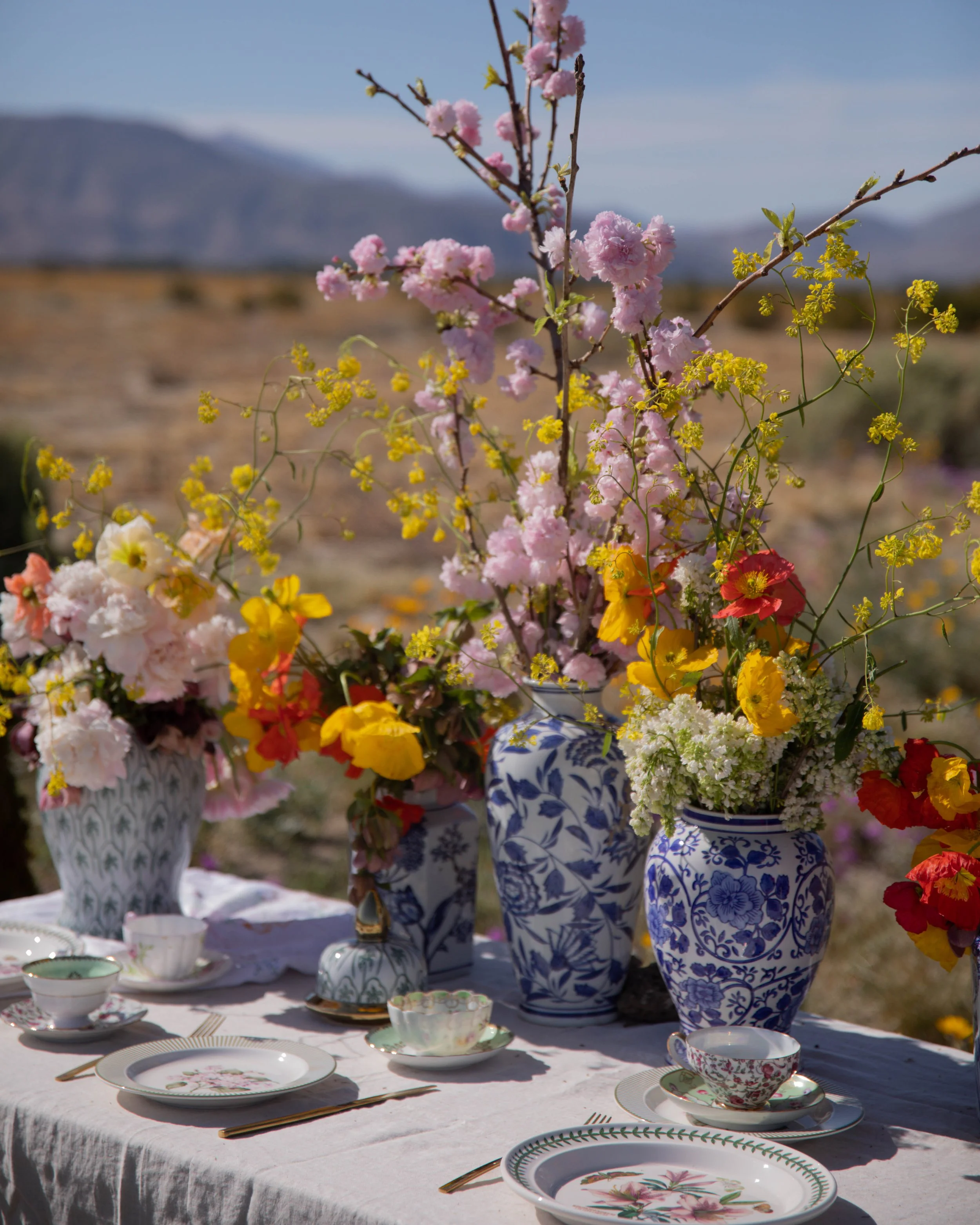 A table set for an outdoor meal with fine china, including teacups and saucers decorated with floral patterns, surrounded by vases filled with pink and yellow flowers, with a scenic mountain landscape in the background.