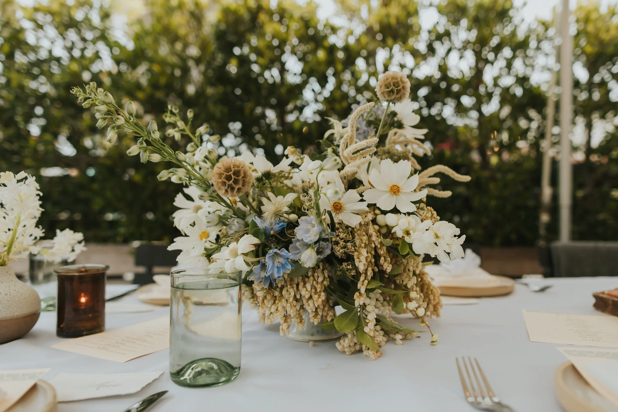 A table decorated with a floral centerpiece, candles, and place settings, set outdoors with trees in the background.