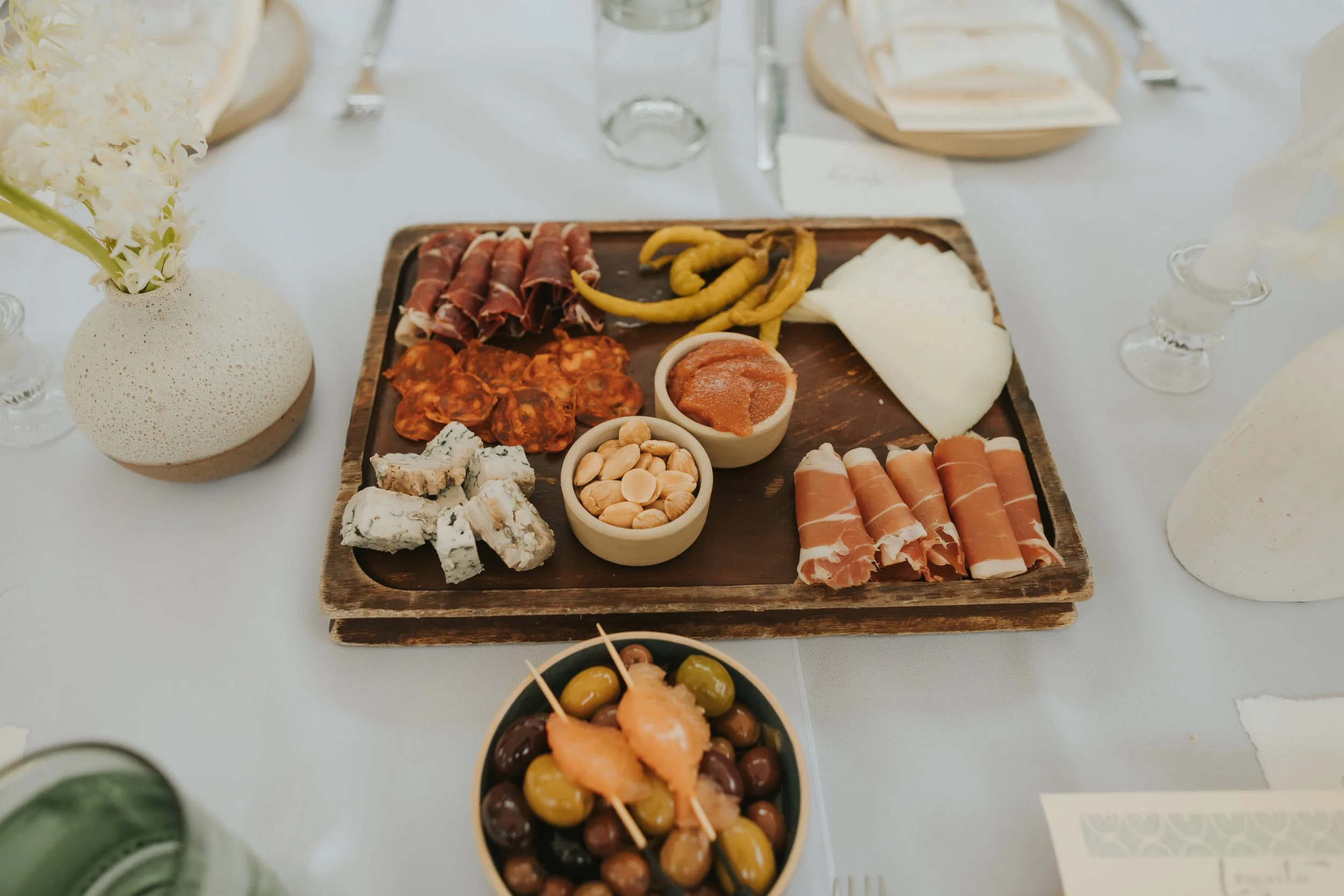 A wooden charcuterie board with slices of cured meats, cheese, marinated red pepper, almonds, and crackers. Surrounding the board are small bowls with olives, cheese, and sauces, on a white table with a floral arrangement in a textured white vase.