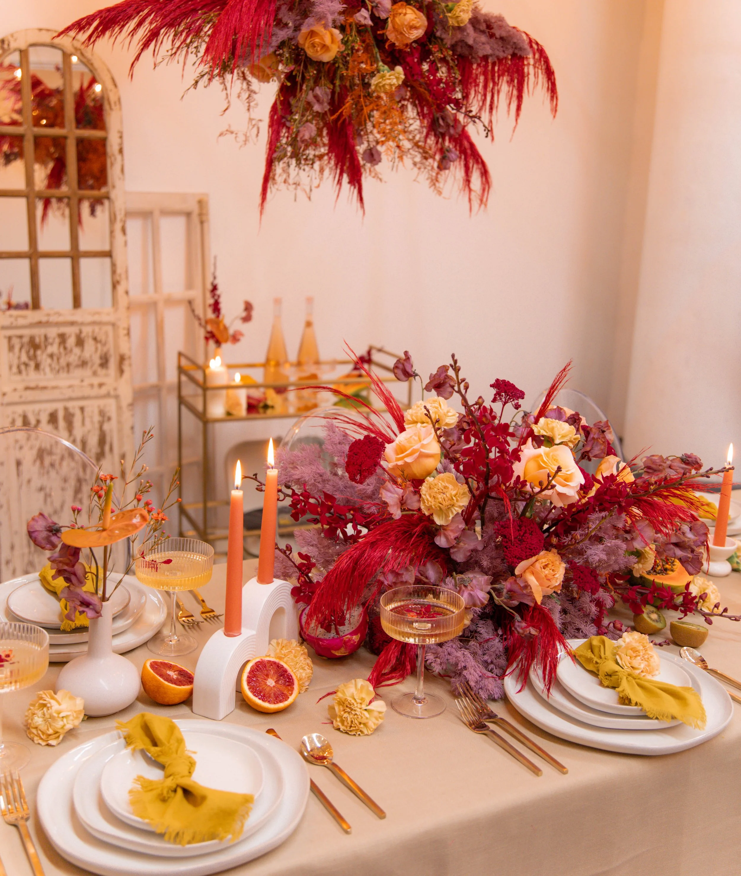 A table setting with white plates, gold flatware, and yellow napkins, decorated with a large floral centerpiece featuring pink, purple, and red flowers and feathers, and topped with hanging pink and purple dried flowers. There are also candles, halve
