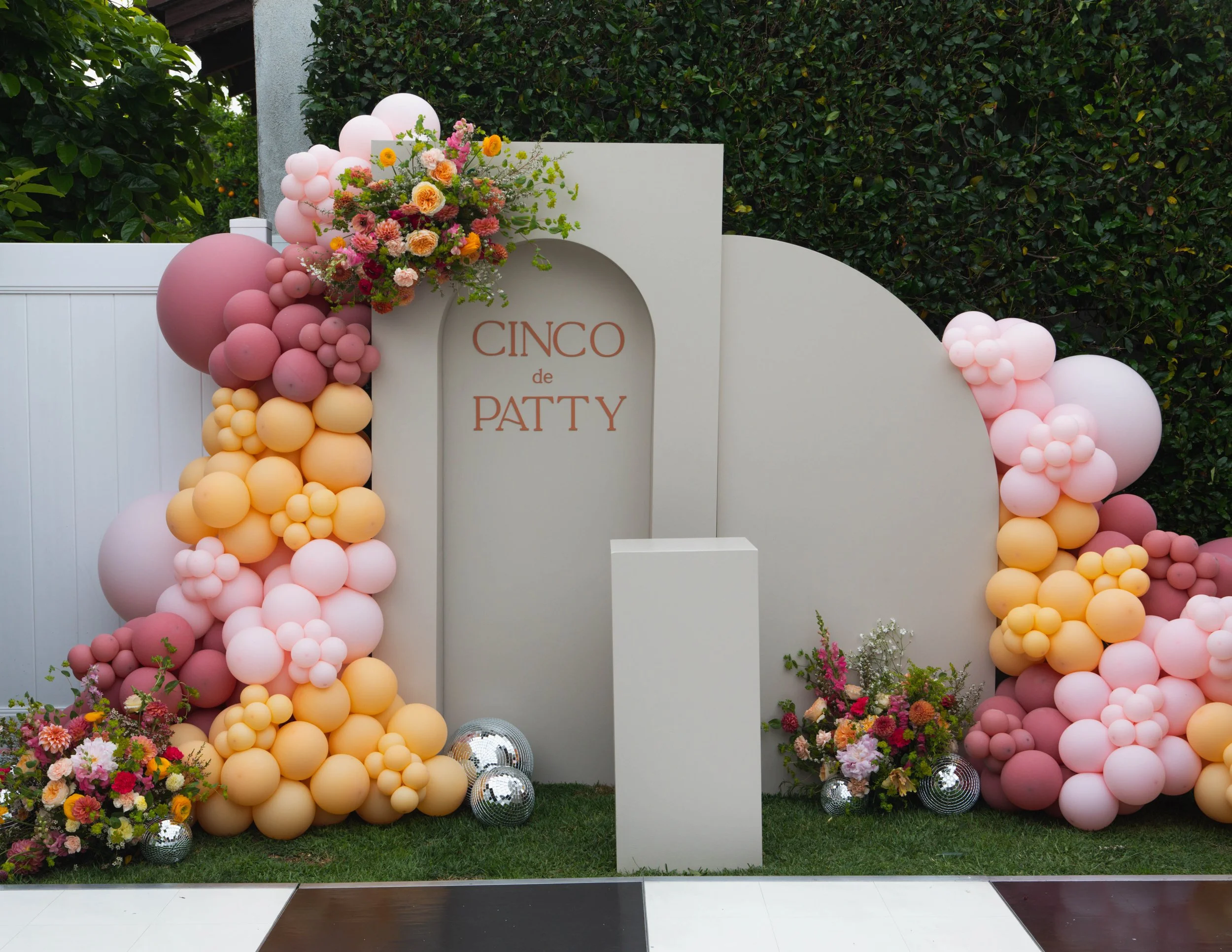 Decorative backdrop with pink and peach balloons, floral arrangements, and silver disco balls, with a sign that reads 'Cinco de Patty'.