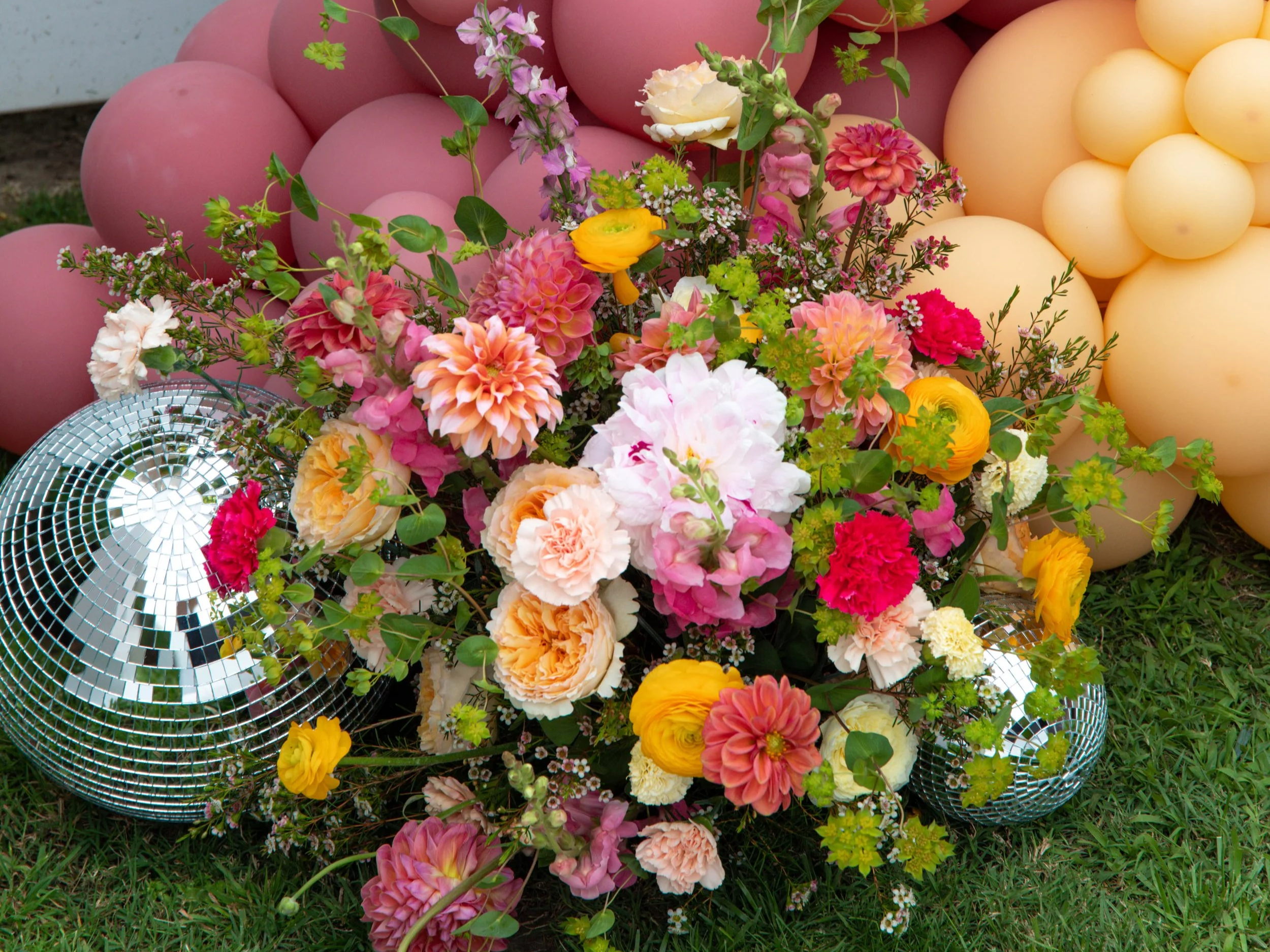 A vibrant bouquet of pink, orange, yellow, white, and purple flowers resting on green grass, surrounded by pink and yellow balloons and a disco ball.