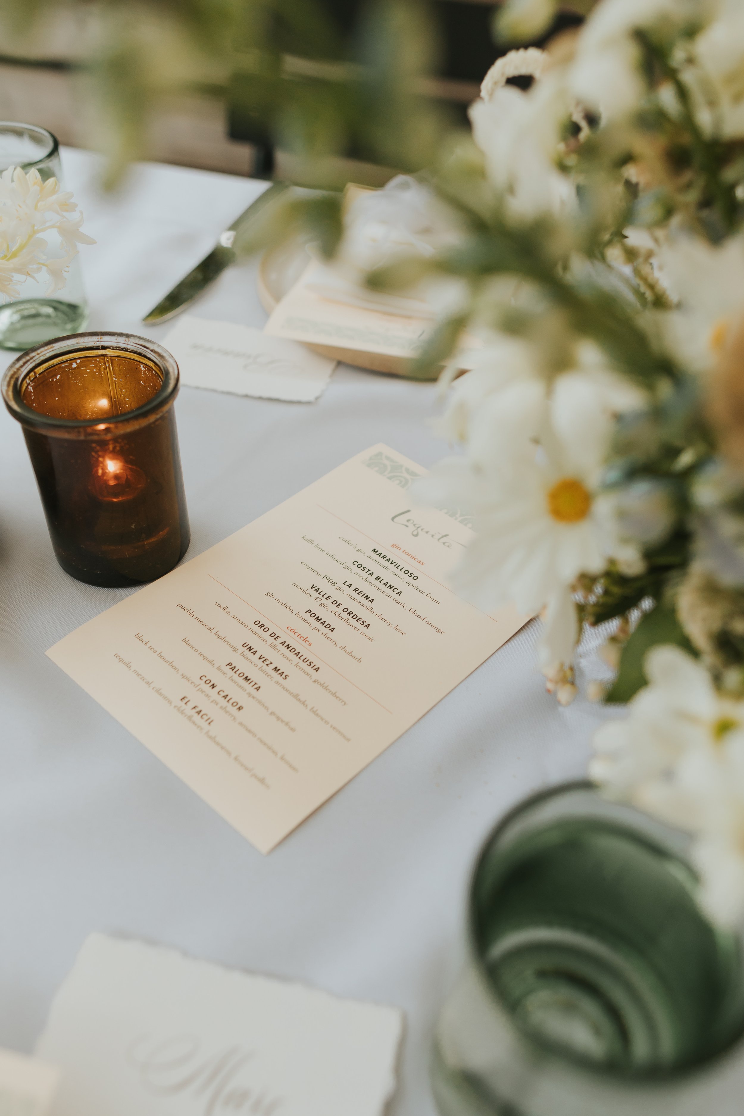 Close-up of a table setup with a menu, a small candle in a brown holder, a glass of water with flower, and floral decorations.