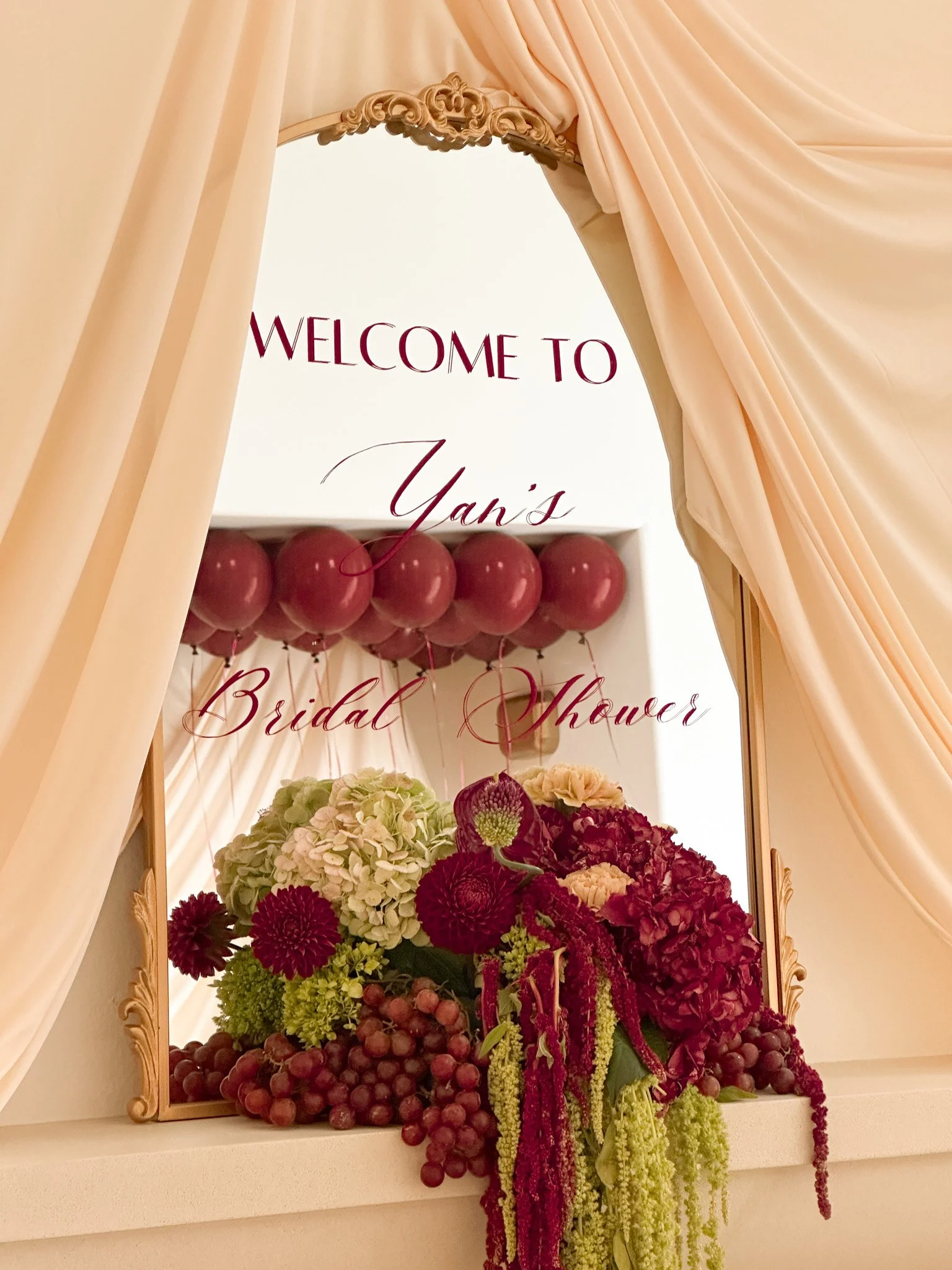 A floral display with a variety of red and green flowers. Red balloons and a sign that reads 'Welcome to Yan's Bridal Shower' are visible through a gold-framed mirror and peach curtains.