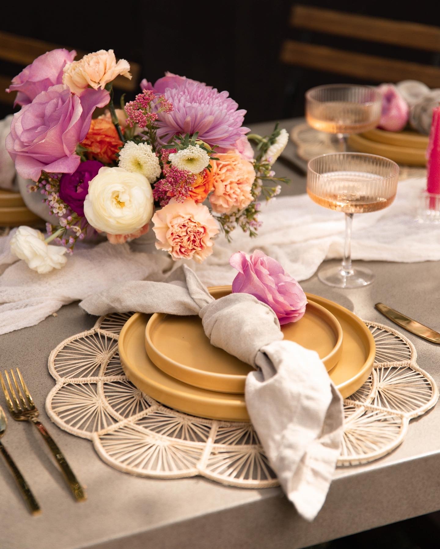 A table setting with a floral centerpiece, pink and white flowers, and a place setting with a yellow plate, a cloth napkin, gold utensils, and decorative doilies.