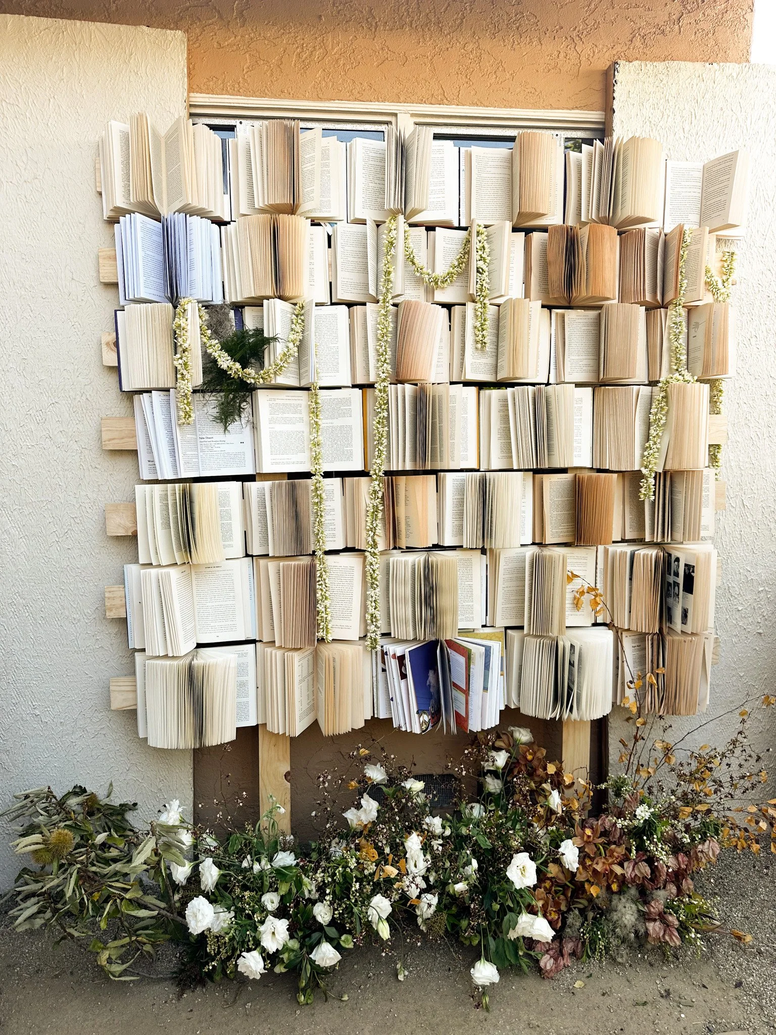An outdoor display of open books arranged in a grid on a wall, decorated with flower garlands, with flowers and plants at the bottom, set against a beige wall.