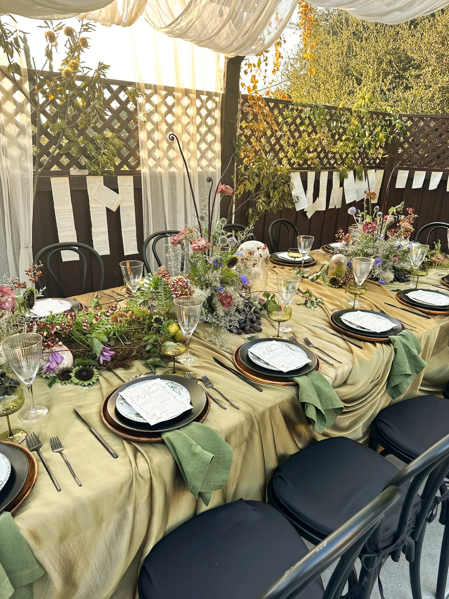 A long dining table decorated with floral arrangements, surrounded by black chairs with green cloth napkins and printed menu cards, set outdoors with a tan tablecloth, with a wooden fence, trees, and draped white fabric overhead.
