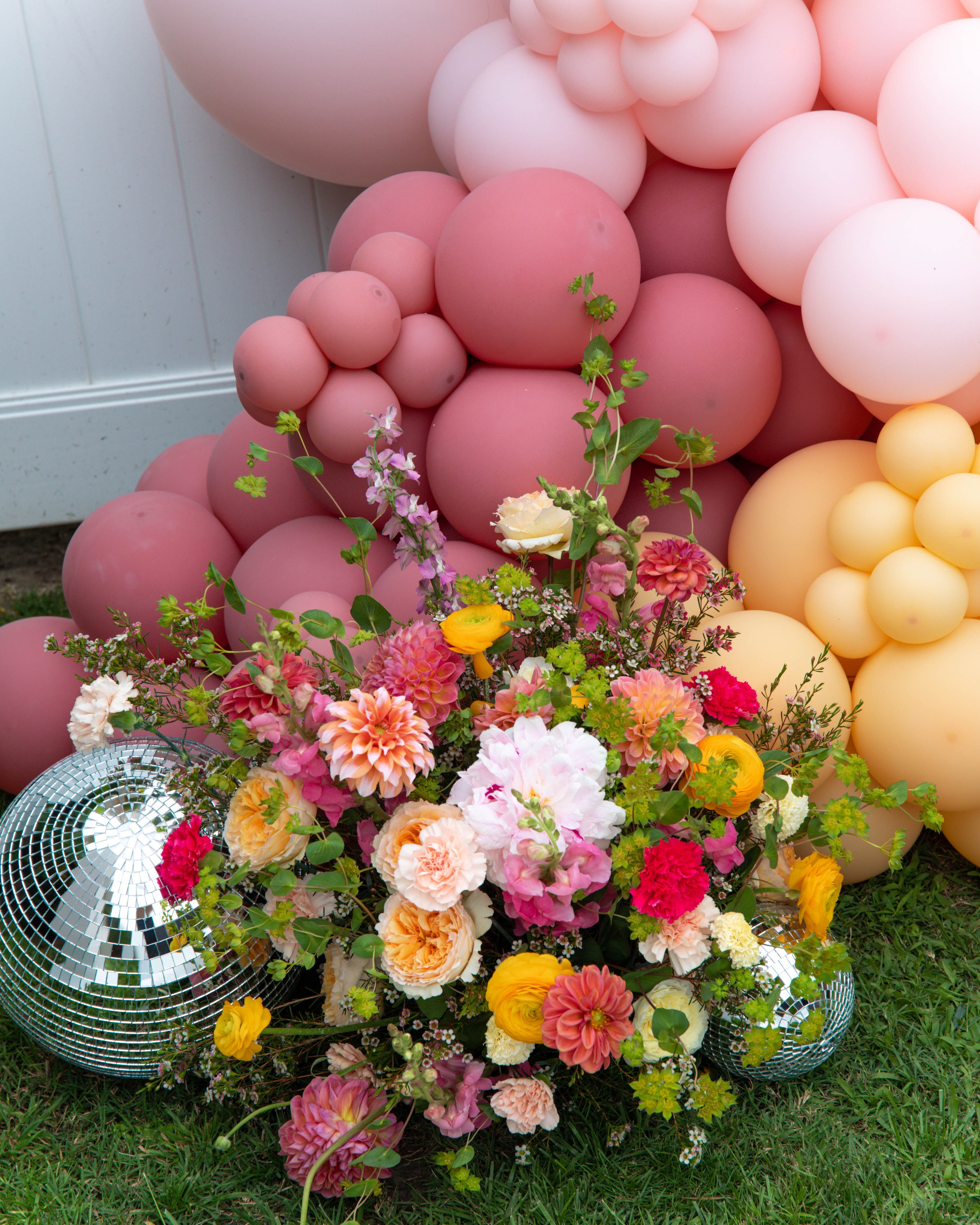 A large arrangement of pastel-colored balloons in pink, yellow, and peach, decorated with fresh flowers, placed outdoors on grass with a white fence in the background.