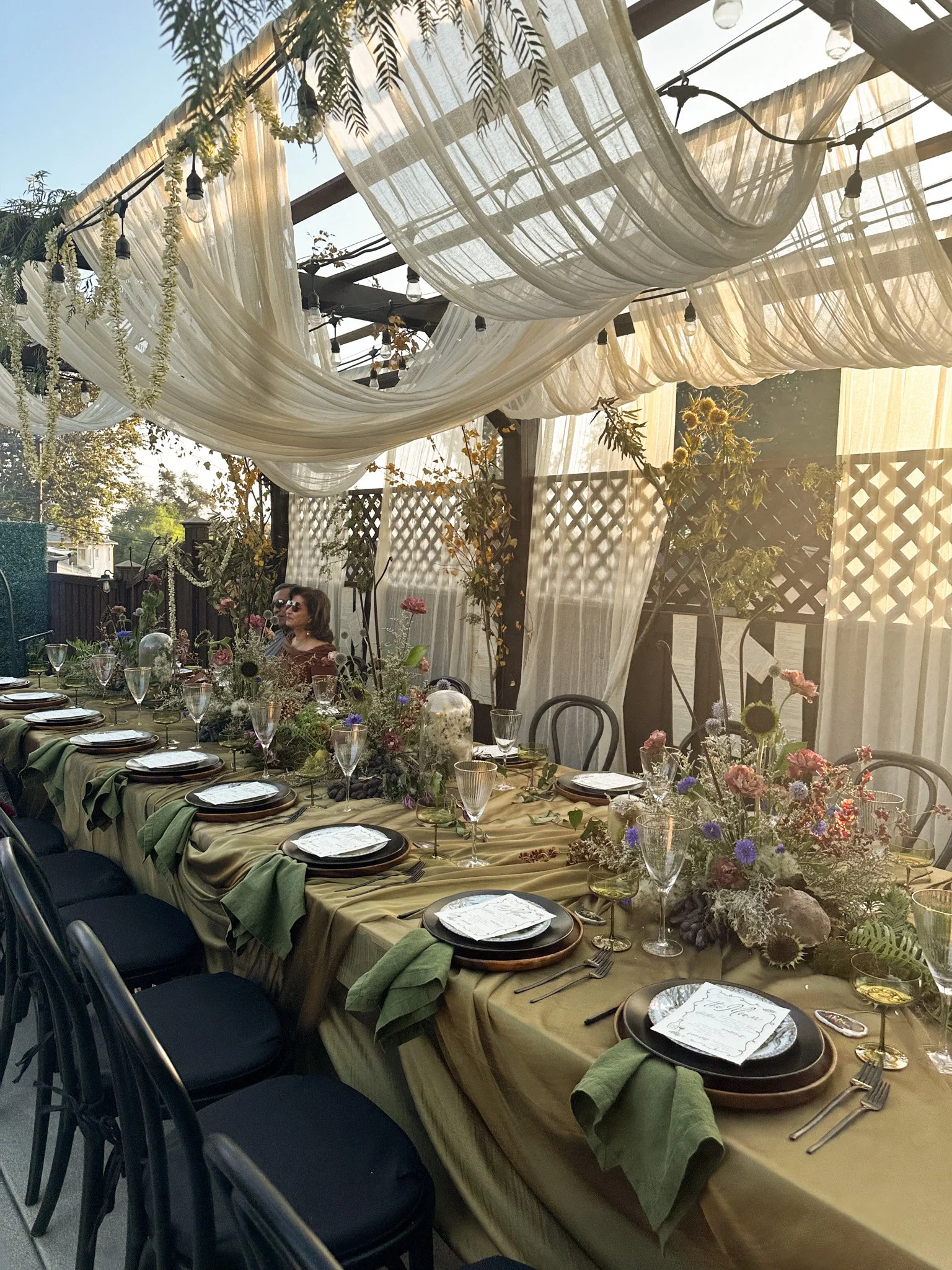 A decorated outdoor dining setup with a long table set with plates, glasses, and floral centerpieces under a canopy of white fabric and string lights.
