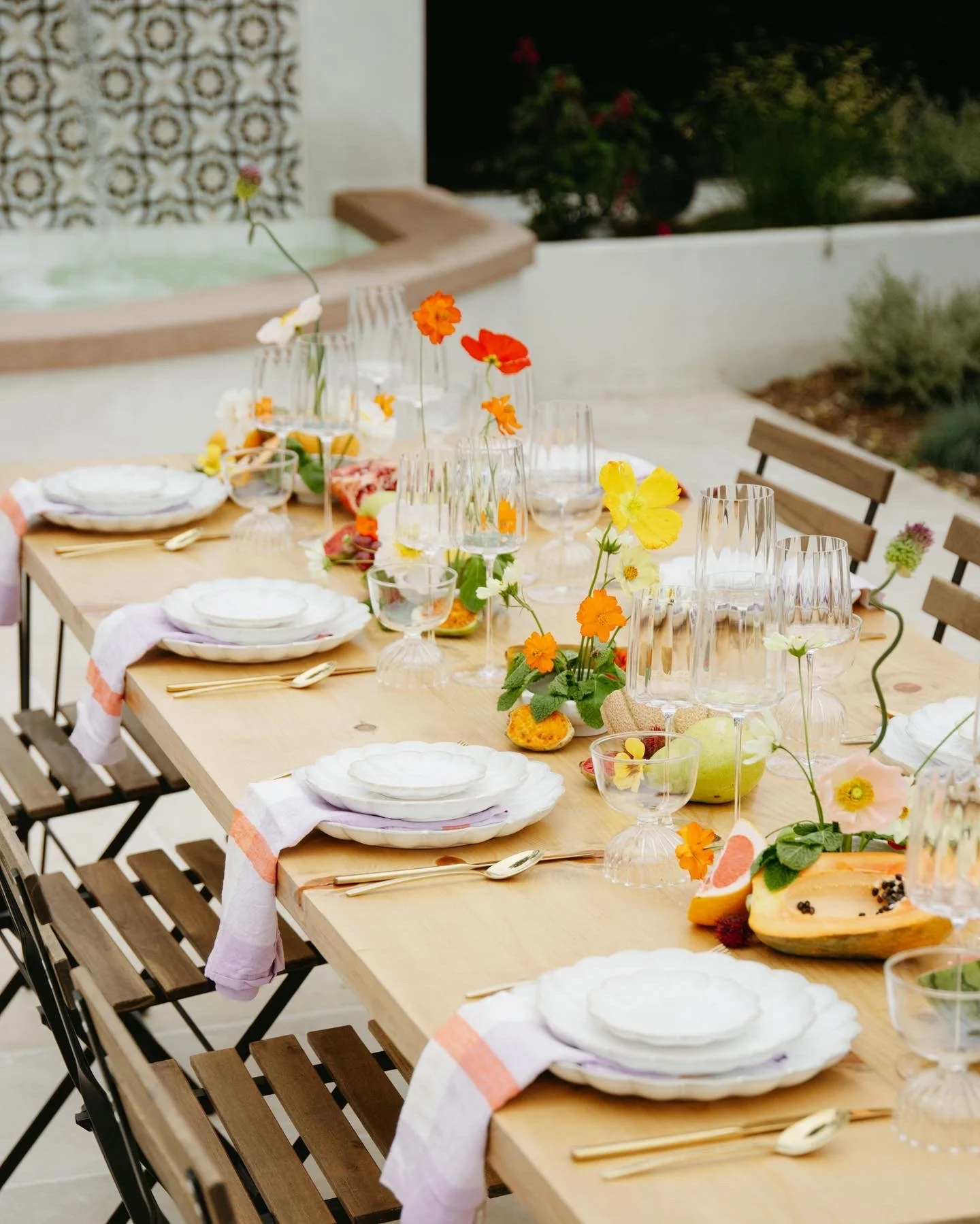 A long outdoor dining table set for a meal with white plates, gold cutlery, glassware, and a centerpiece of colorful flowers including orange, yellow, pink, and white blooms. The table is decorated with cloth napkins with orange edges and has a rusti