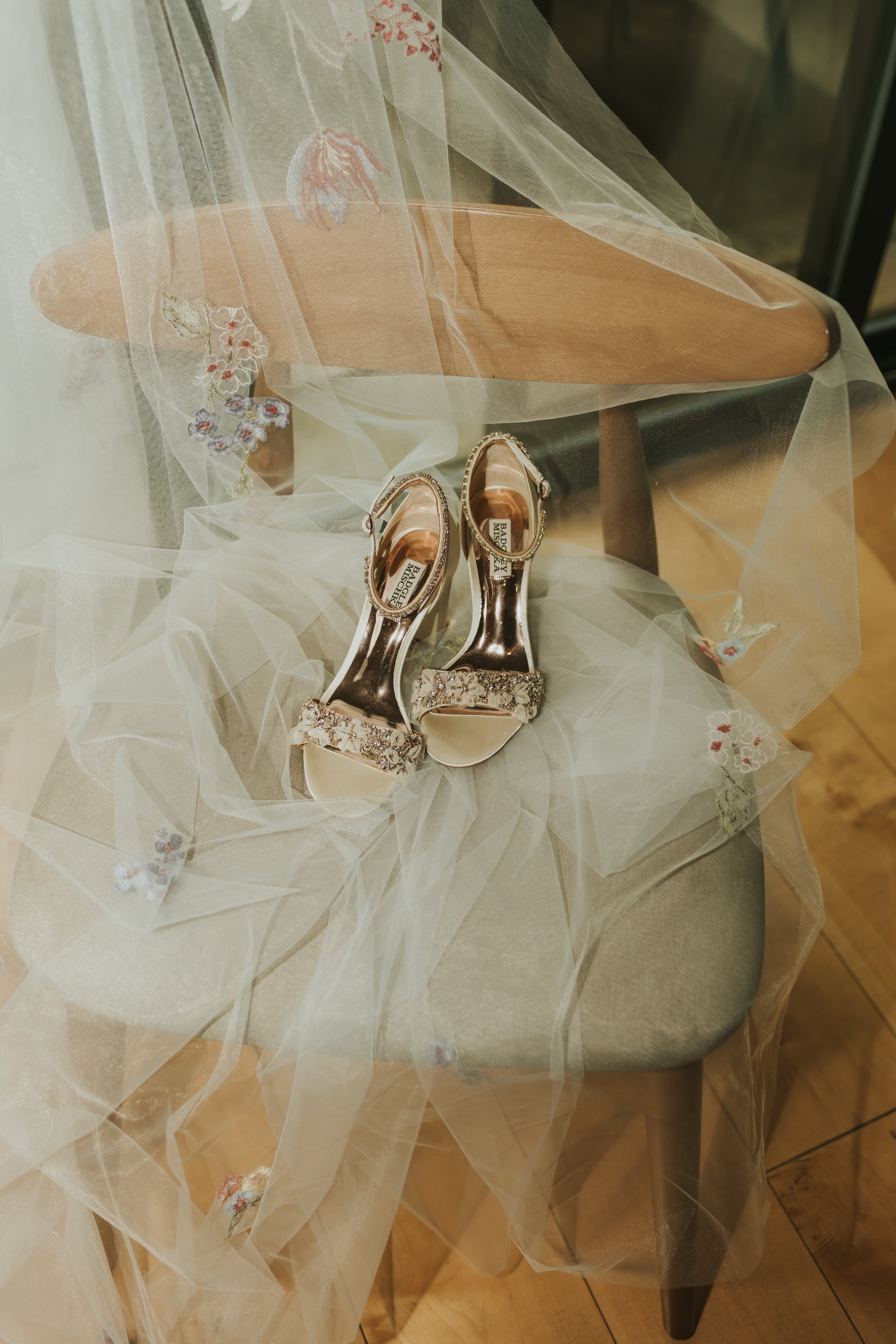 A pair of silver high-heeled shoes with rhinestone embellishments sitting on a chair, covered by sheer, embroidered fabric with floral patterns.