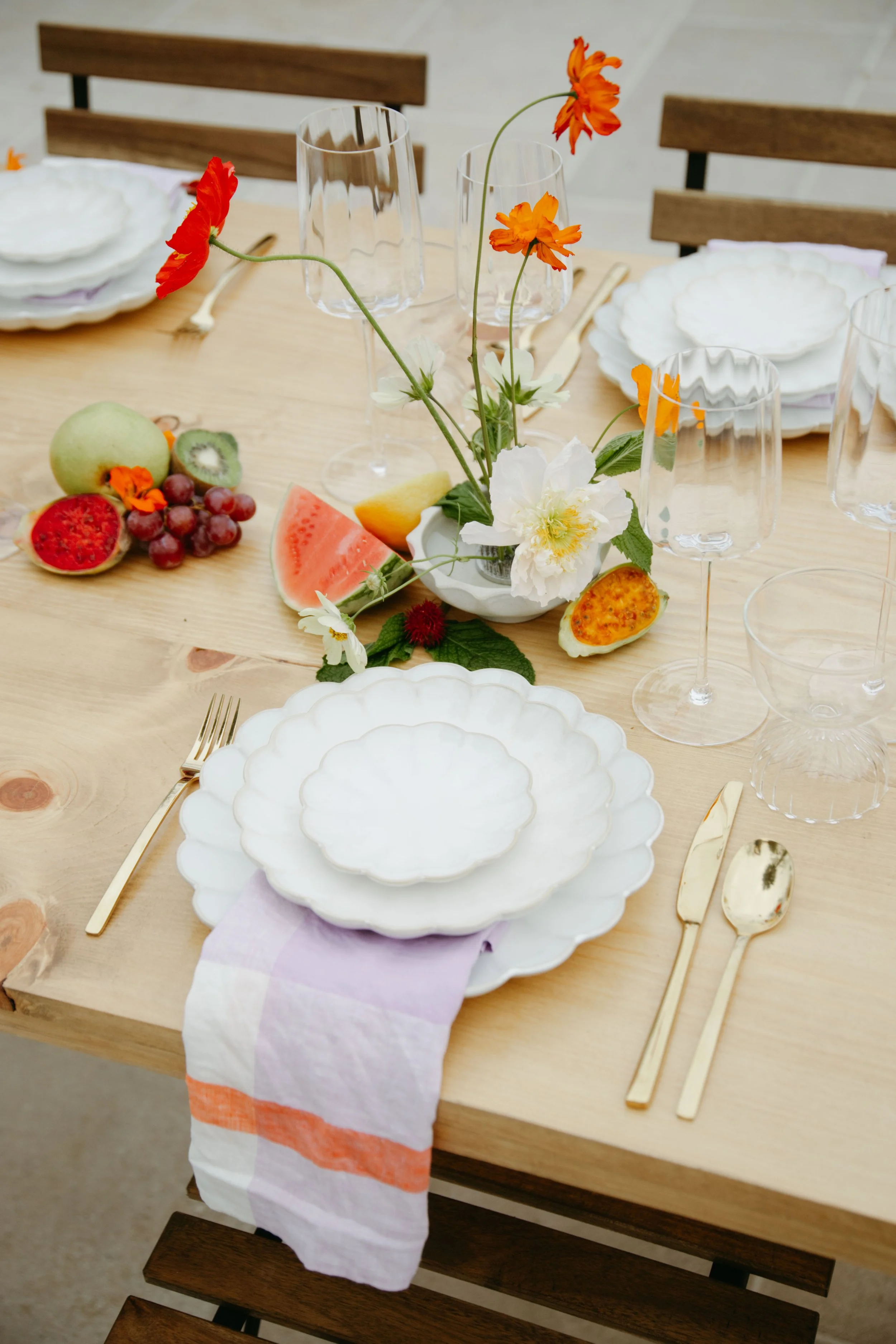 A wooden dining table set with white scalloped dishes, gold flatware, clear glassware, and a floral centerpiece with orange, white, and yellow flowers. Various fruits like kiwi, watermelon, grapes, and apple are placed on the table. The table is surr