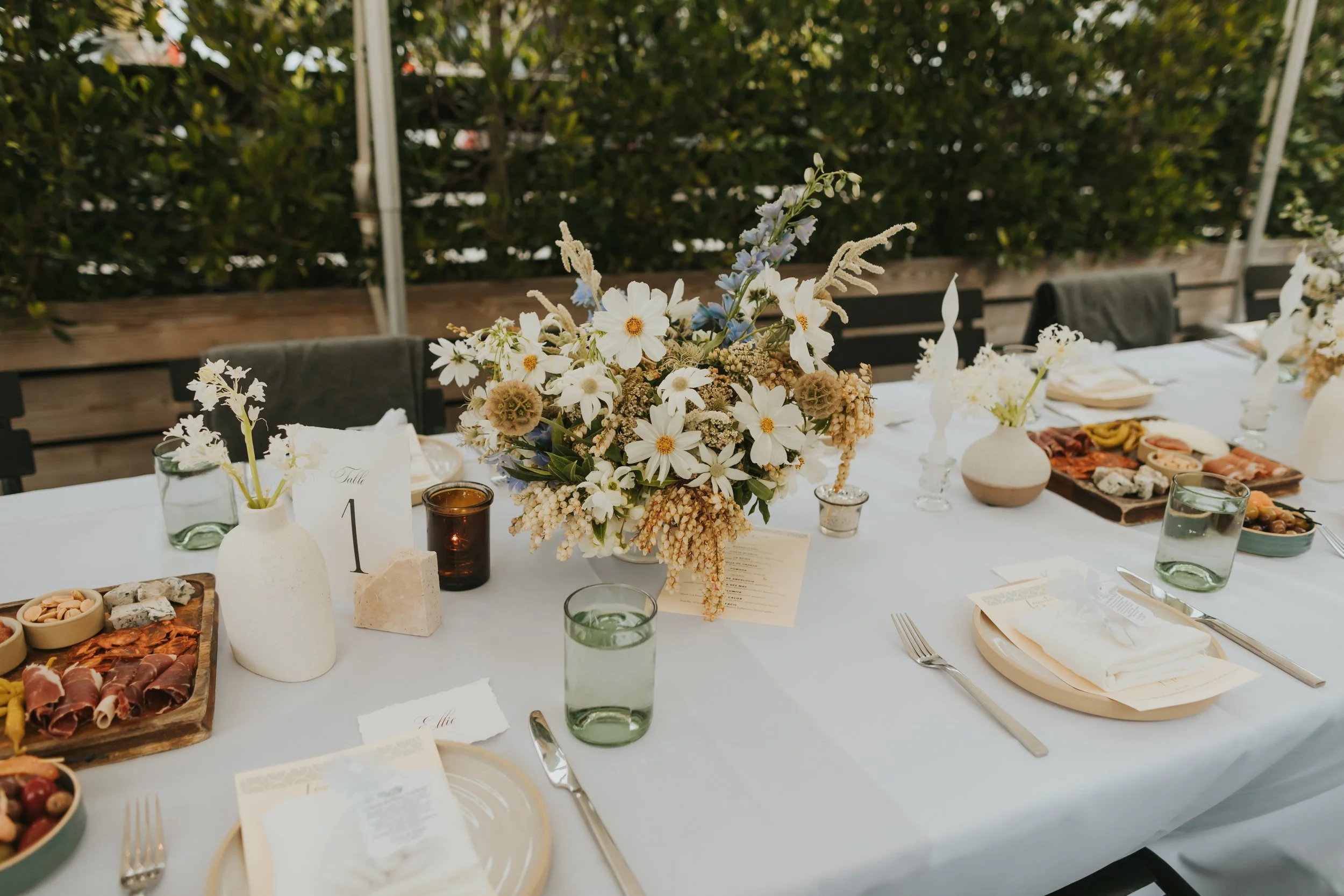 A white table with a floral centerpiece consisting of white daisies, blue delphiniums, and other flowers. The table is set with plates, forks, knives, water glasses, and small bowls of snacks. A small white card with the number 1 is placed on the tab