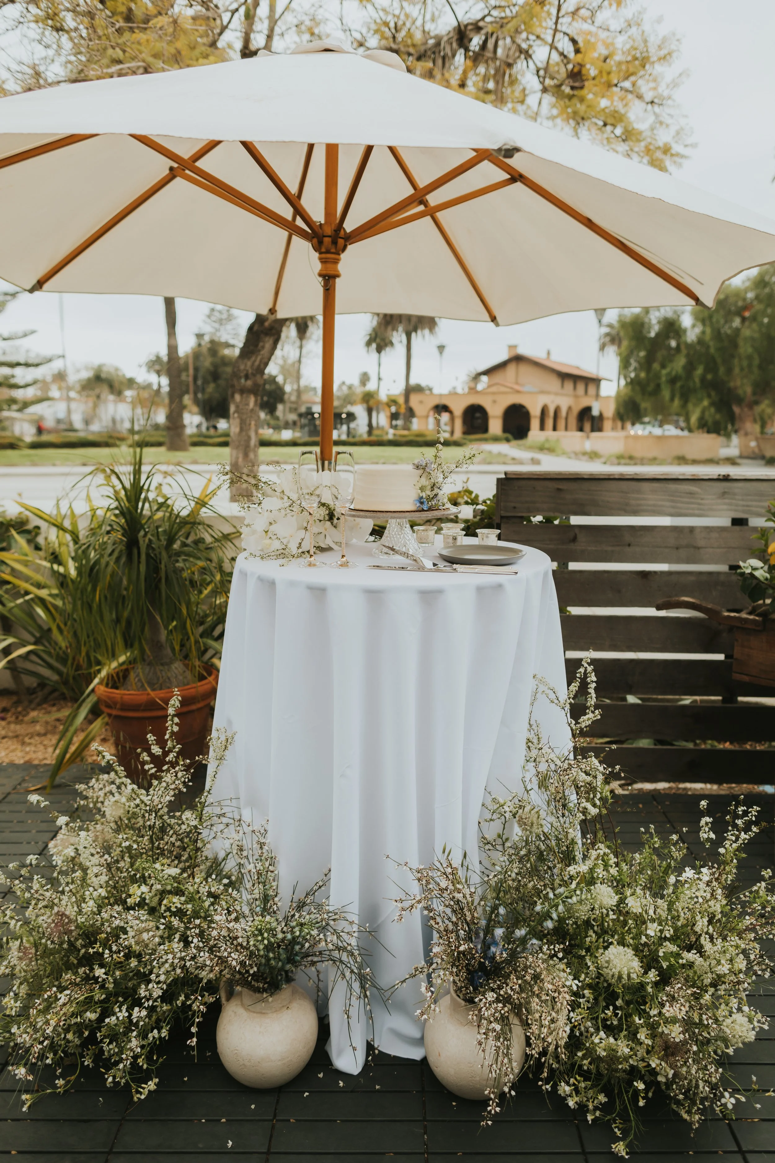 A table with a white tablecloth under a large white umbrella, decorated with flowers and glassware, surrounded by potted plants and white vases with flowers, set outdoors in a garden area with trees and a building in the background.