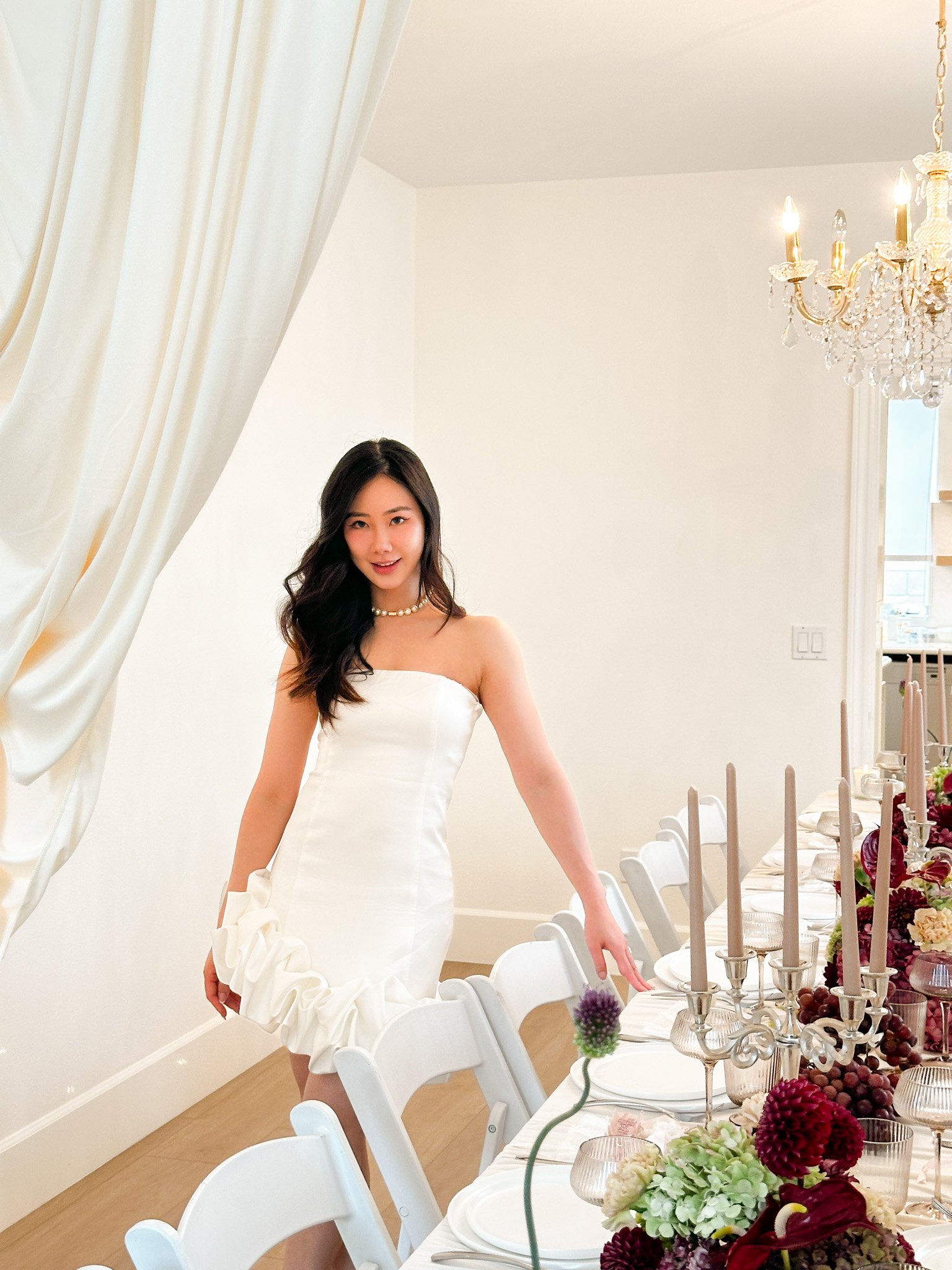 A woman in a white strapless dress with long dark hair and a pearl necklace standing next to a dining table decorated with flowers, candles, and tableware in a well-lit room.