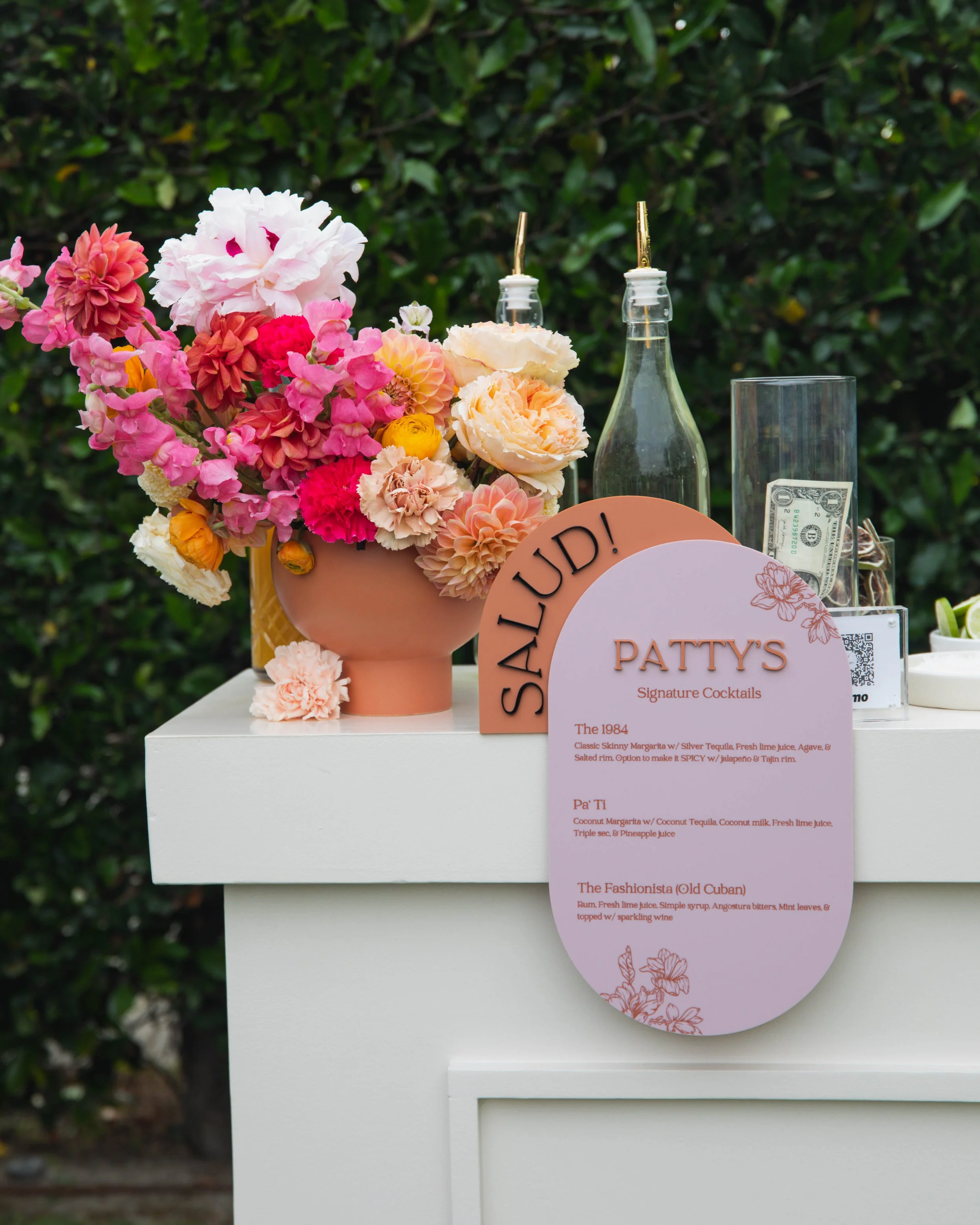 A white table with a pink floral arrangement in a vase, a glass container with a dollar bill, and a menu card titled 'Patty's Signature Cocktails' listing three drinks with ingredients. The background consists of green foliage.