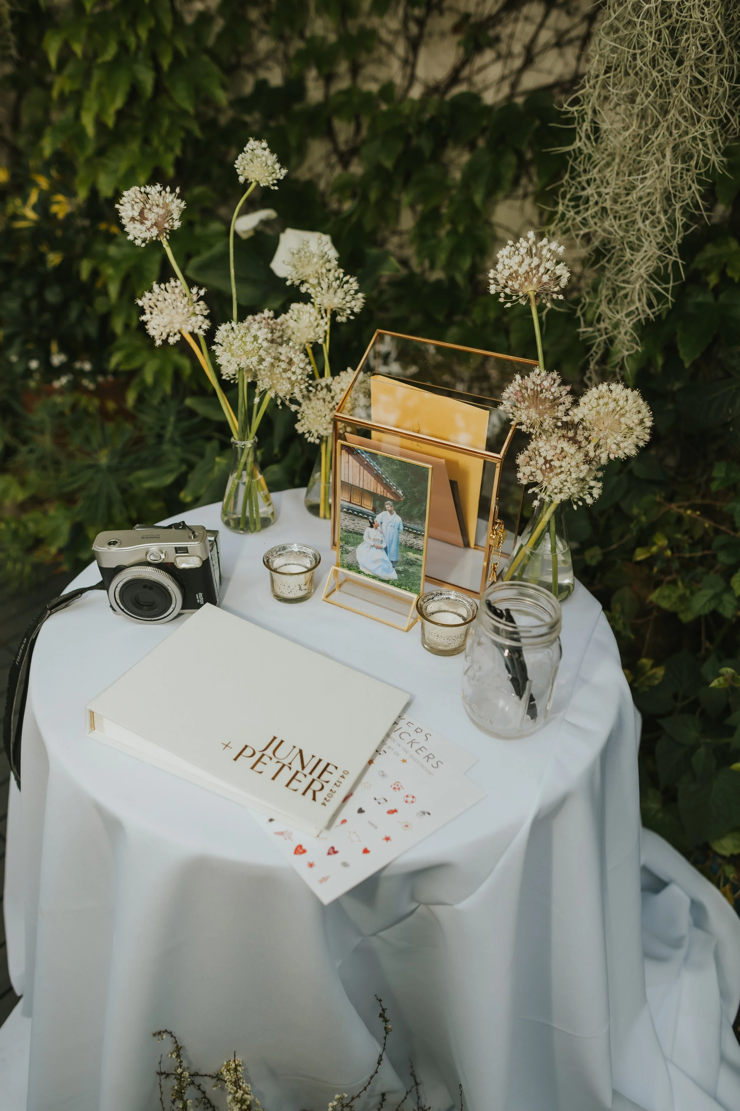 A round table covered with a white cloth, decorated with two vases of white flowers, picture frames, a vintage camera, candles, and wedding-related materials including a book titled 'JUNE + PETER', wedding invitation cards, and a pen jar, outdoor set