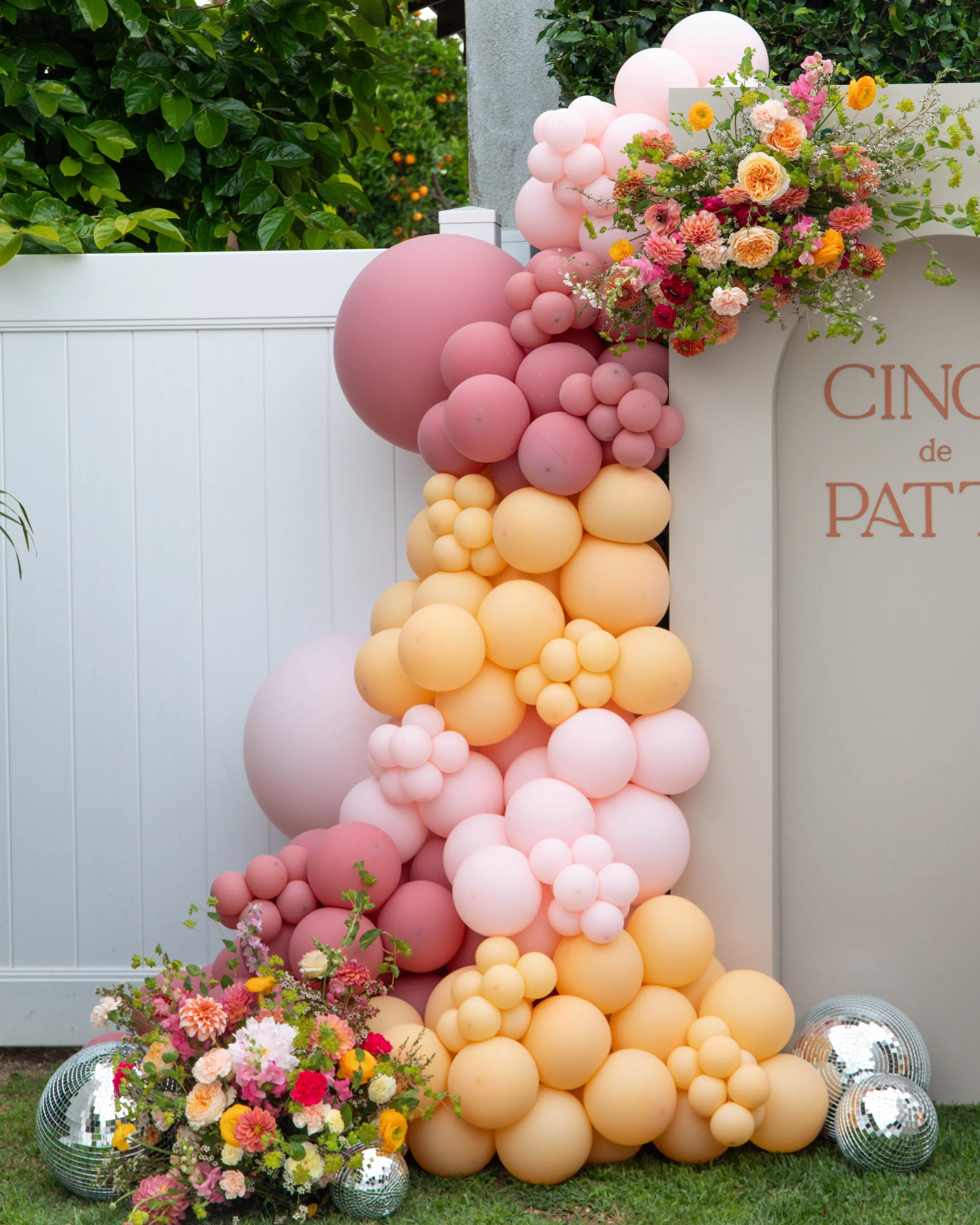 Decorative arrangement with pink, yellow, and peach balloons and a colorful flower bouquet positioned on the ground, with some small disco balls placed among the flowers for decoration.