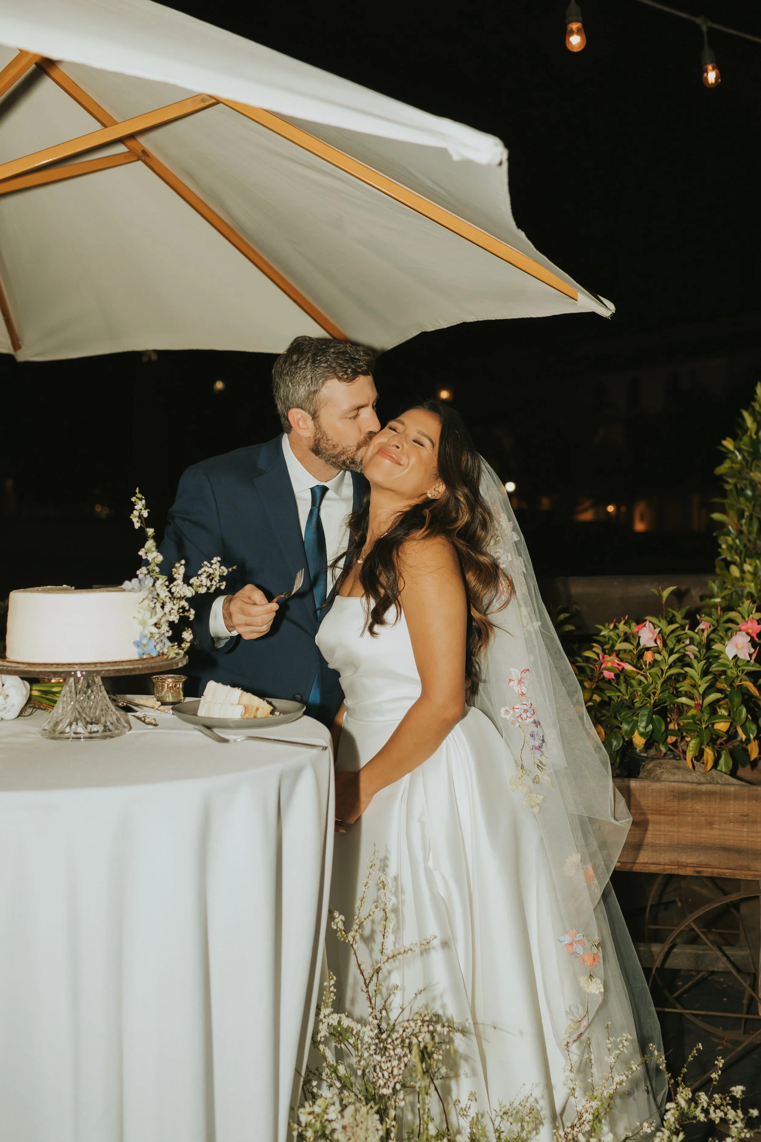 A newlywed couple sharing a kiss at their wedding reception, with the groom in a navy suit and the bride in a white wedding gown, standing near a table with cake and flowers under a large umbrella at night.