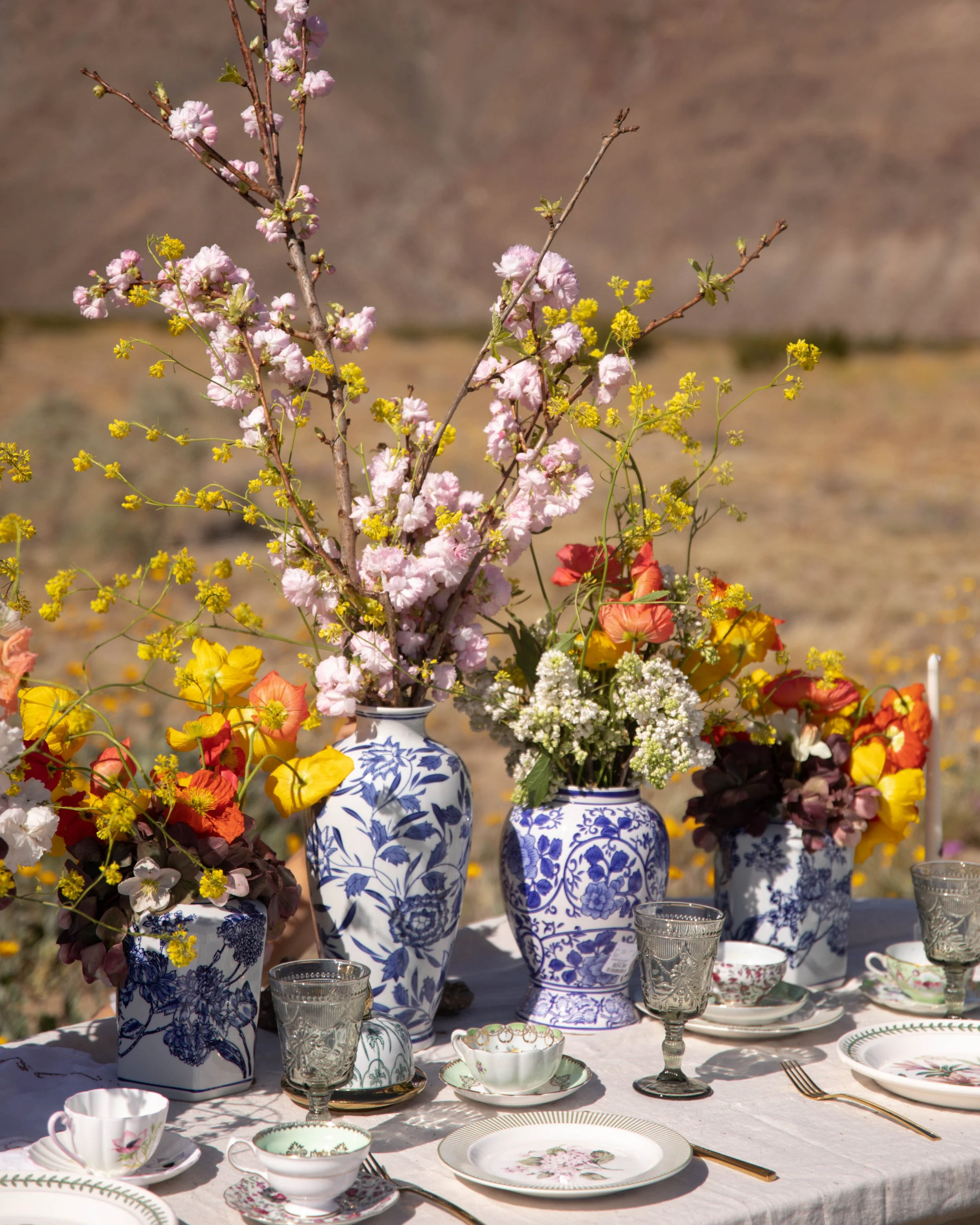 Table with white tablecloth, floral china dishes, glassware, and silverware, decorated with a large bouquet of pink, yellow, and white flowers in blue and white vases. Outdoor setting with blurred background.