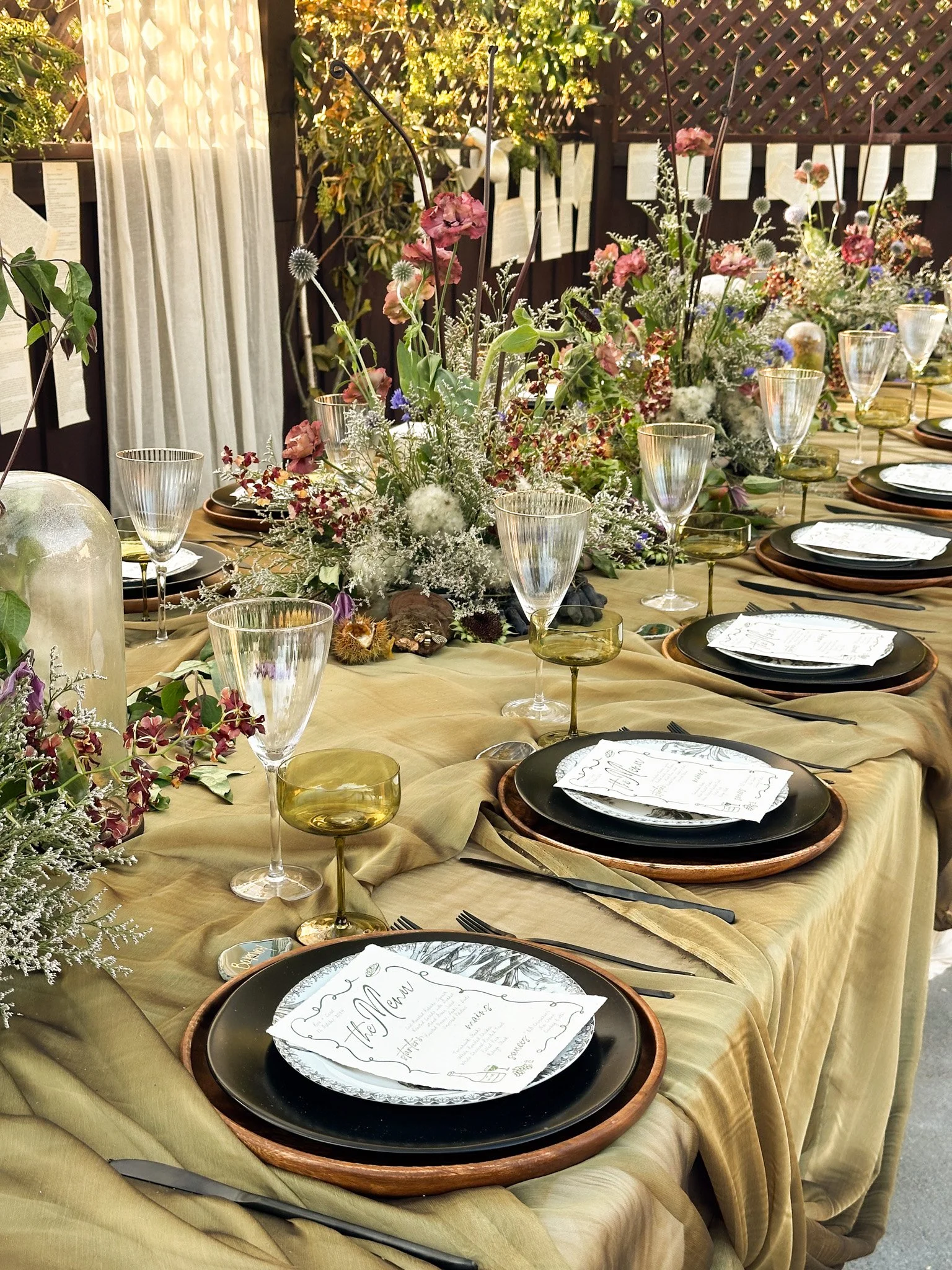Elegant dining table set with black plates, white menus, clear wine glasses, and gold-tinted drinking glasses, decorated with a lush arrangement of pink, white, and purple flowers on a golden tablecloth, outdoors with a decorative background.